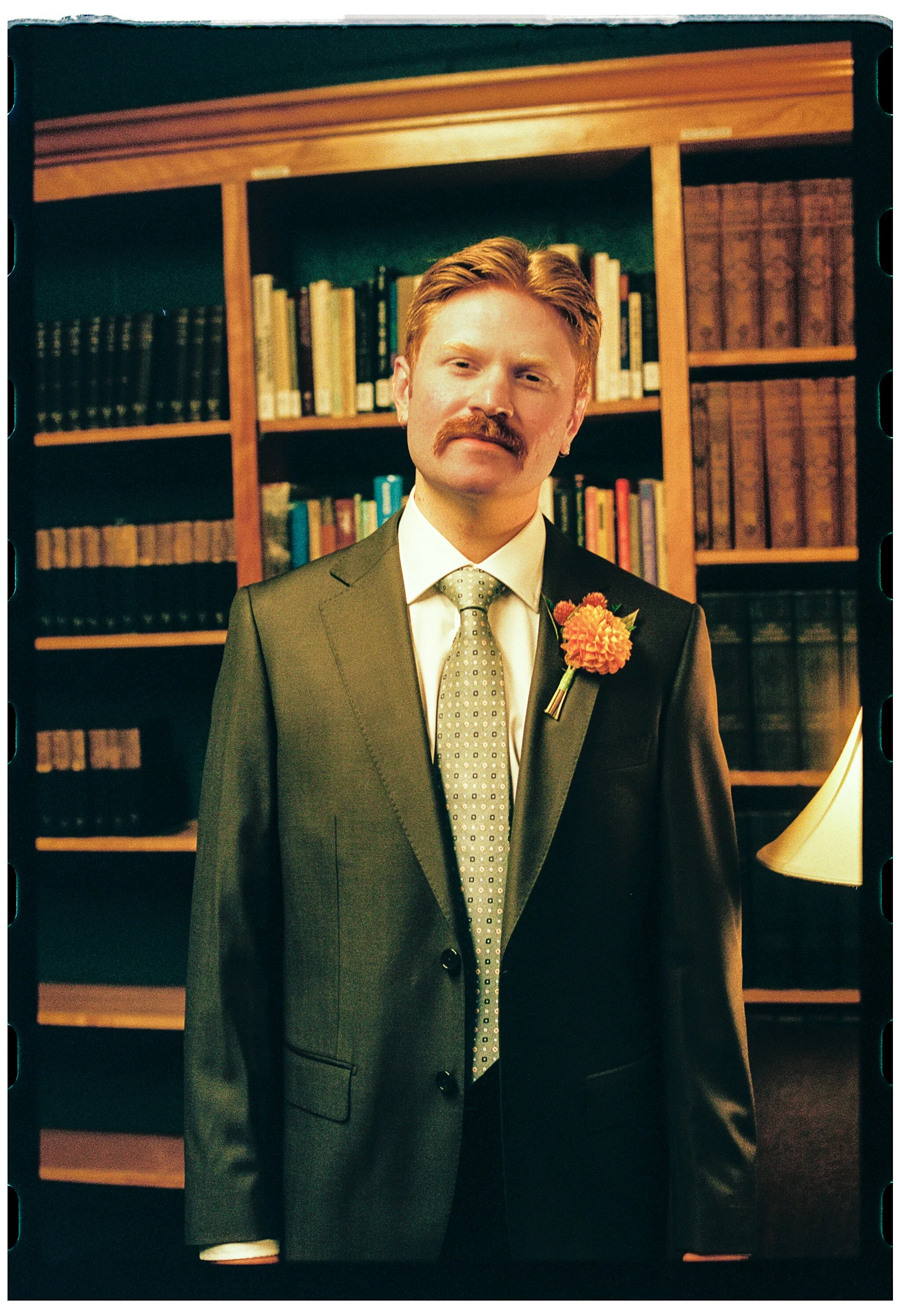 A man with red hair and a mustache in a suit with a flower on his lapel, standing in front of a bookshelf filled with books.
