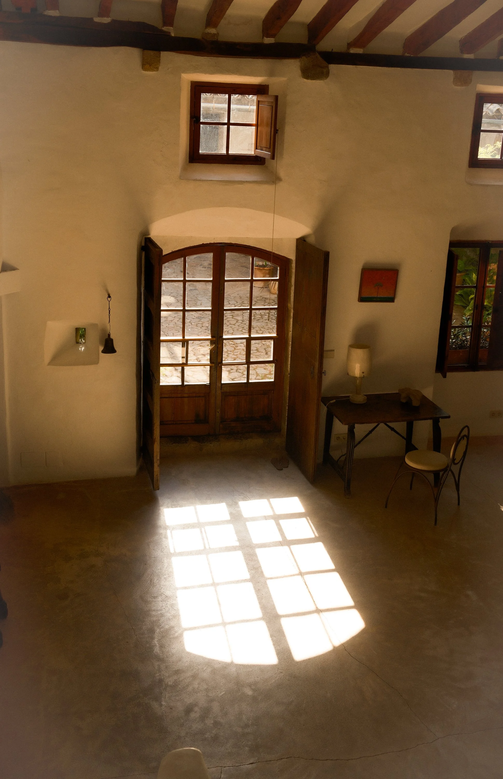 Interior view of a room with sunlight streaming through wooden doors and windows, casting grid-like shadows on the floor. The room has white textured walls, wooden beams on the ceiling, small decorative items, and furniture, including a small table w