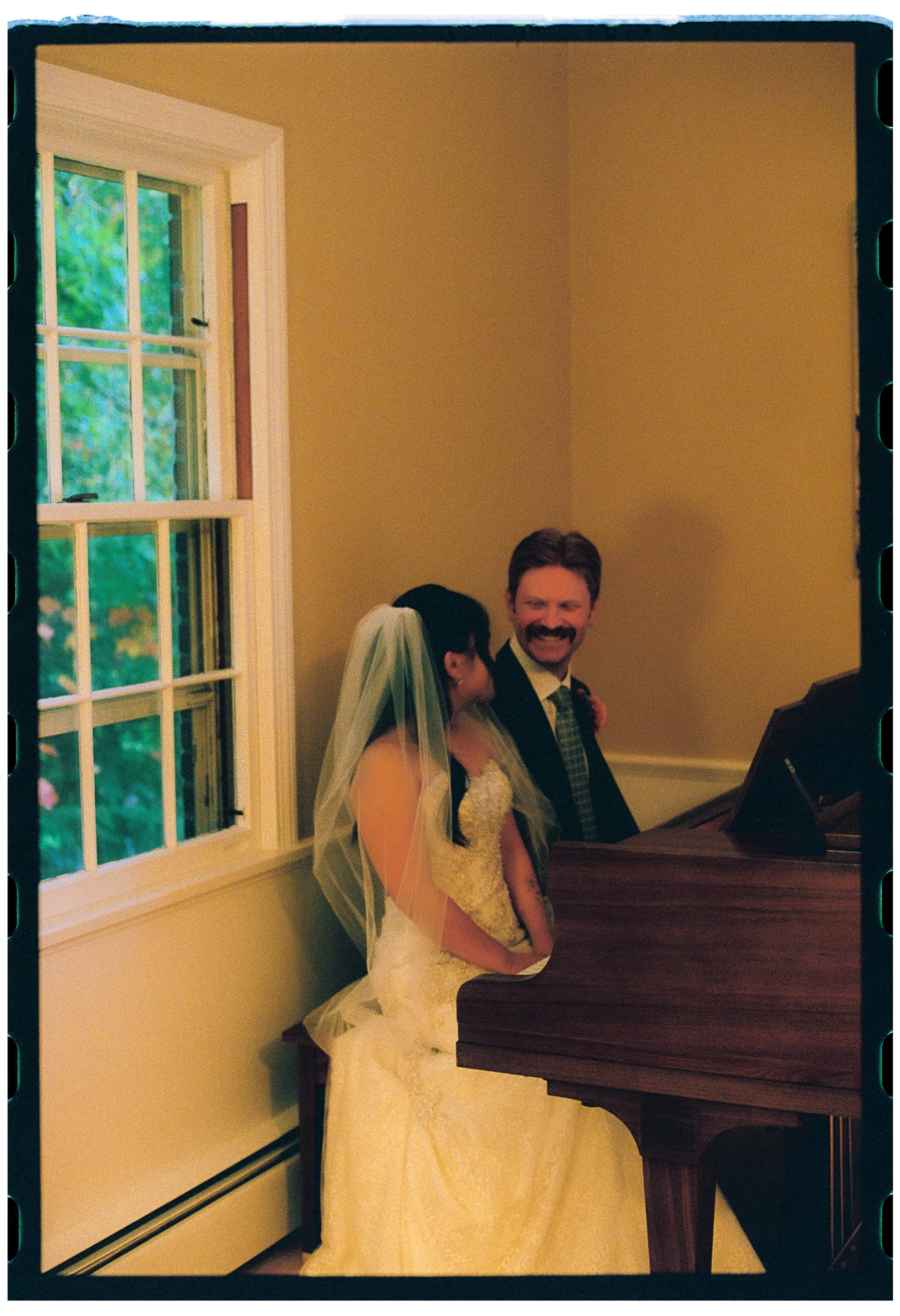 A bride and groom standing together near a window and a piano in a warmly lit room during a wedding celebration.