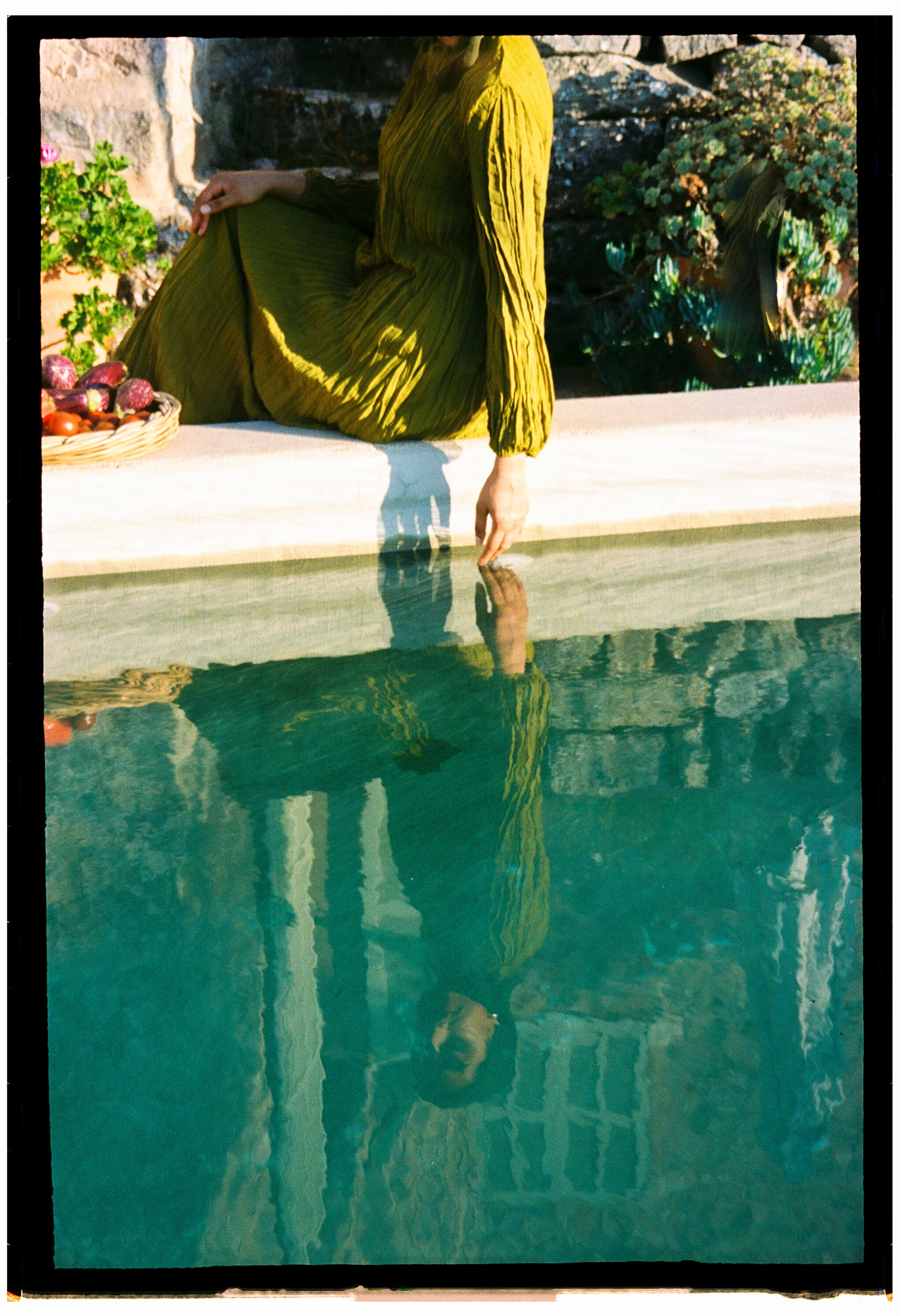 A woman in a green pleated dress sitting on a ledge beside a pool, with her reflection visible in the water. The background includes rocks and greenery. destination wedding nikkah Shot on 35mm film