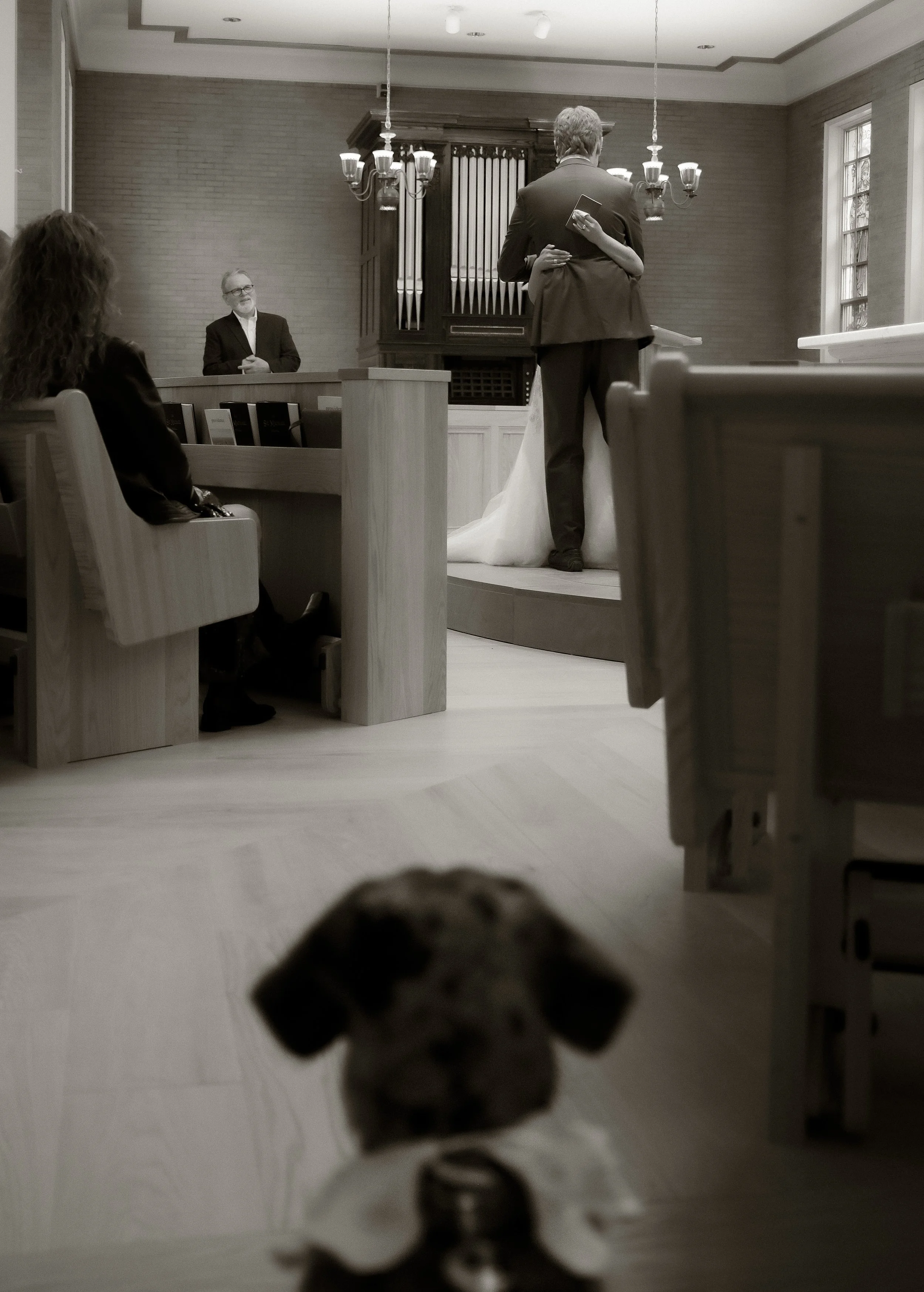 A small puppy looking at a wedding ceremony scene shown through the perspective of the puppy, with the couple hugging in front of an organ.