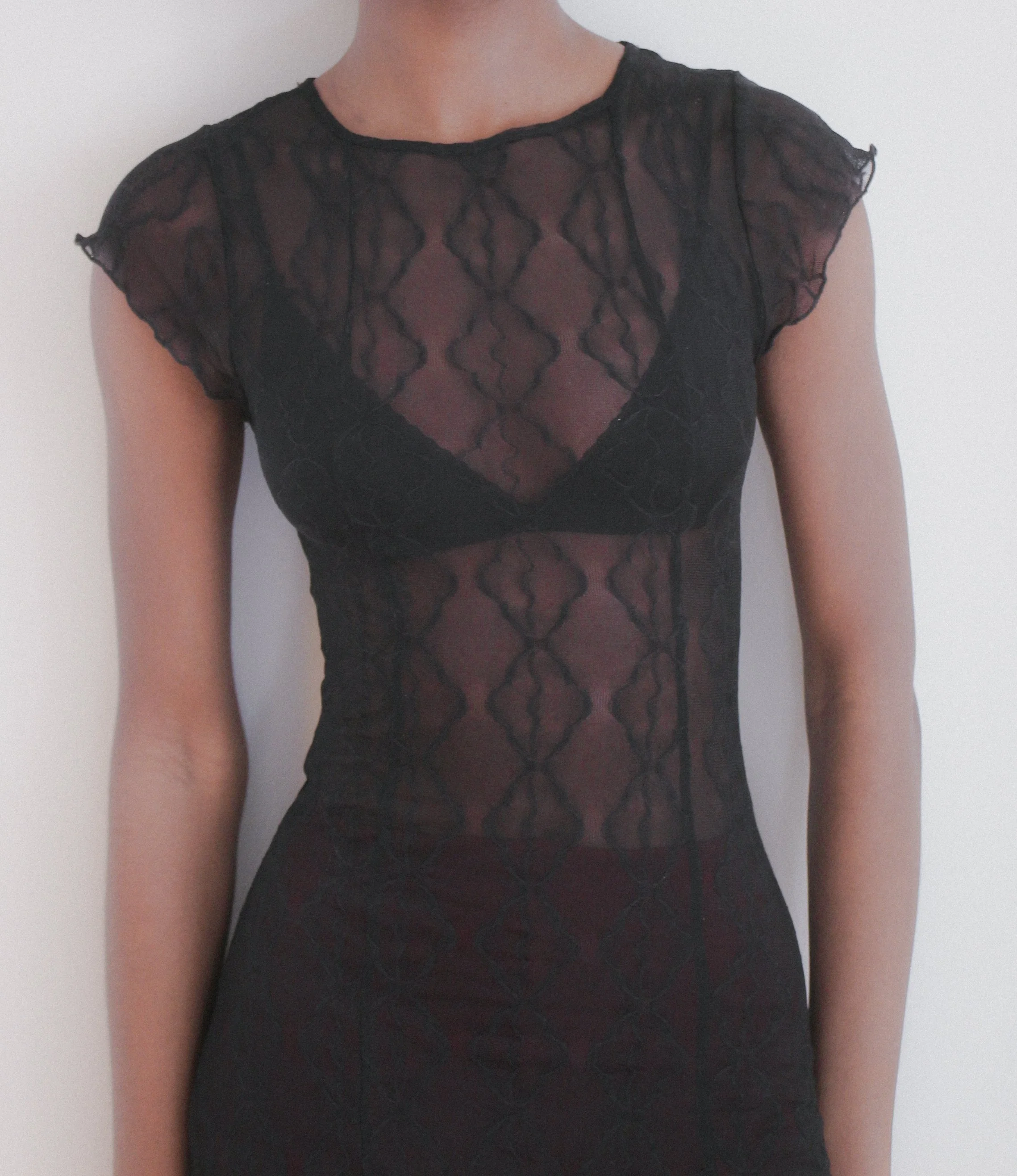 Close-up of a person wearing a black sheer lace top with a geometric pattern, showing their shoulders and part of their neck against a plain white background.