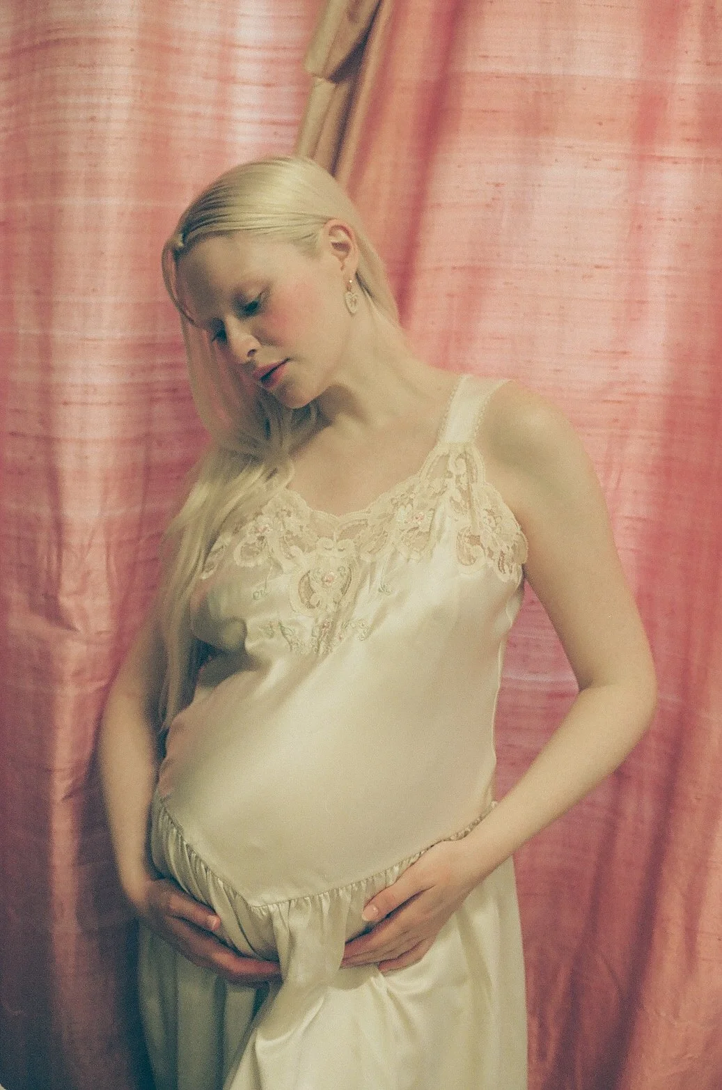 A pregnant woman with long blonde hair looking down while holding her belly, standing in front of pink curtains.