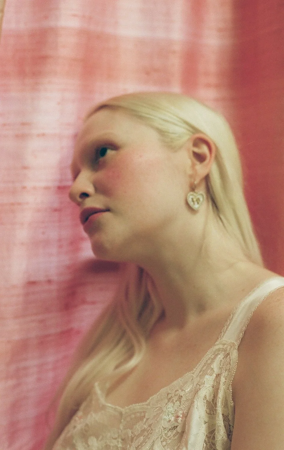 Close-up of a young woman with long blonde hair, wearing heart-shaped earrings and a lace top, with a pink textured background.