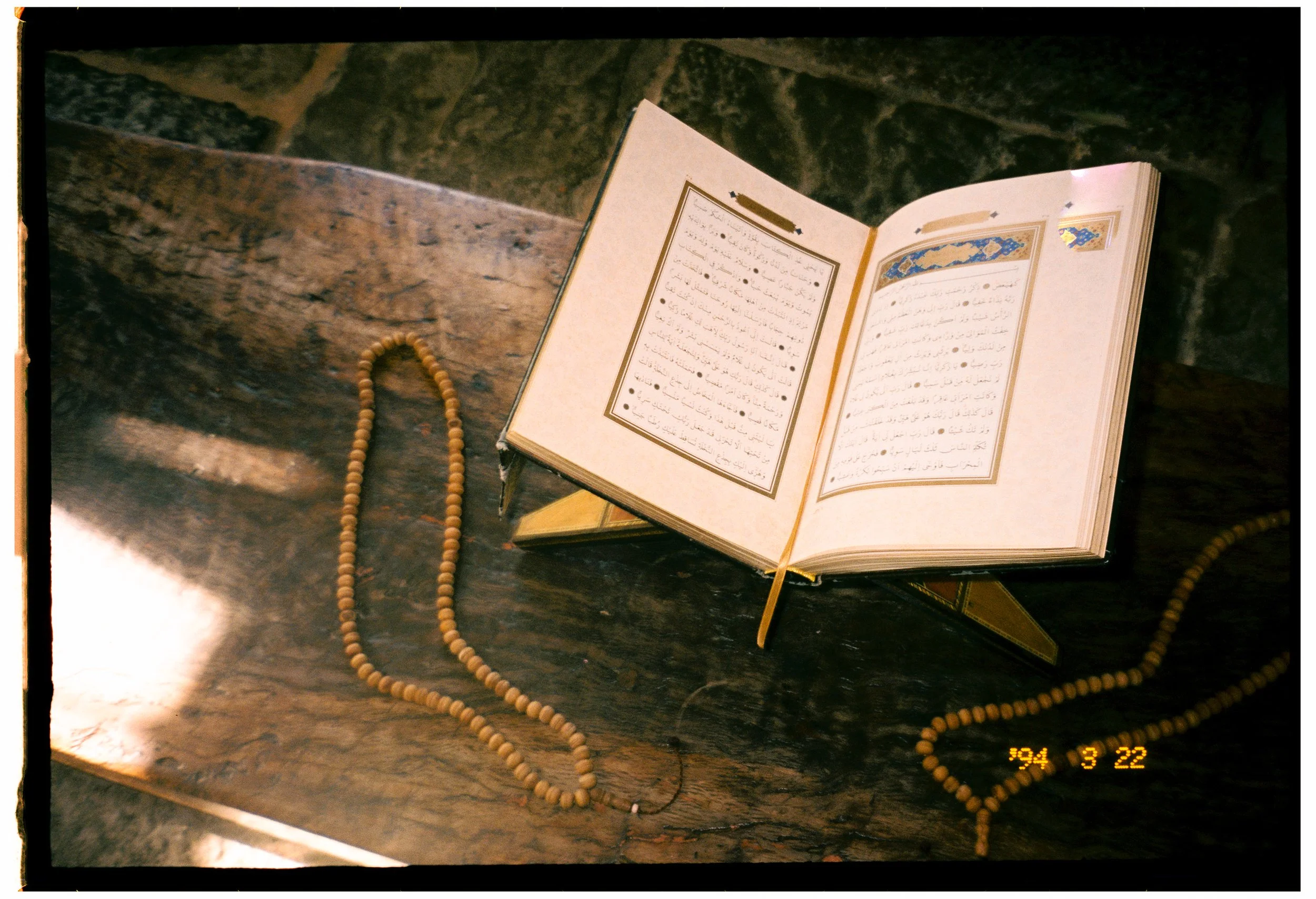 An open book with Arabic text and decorative borders, resting on a wooden surface. A string of prayer beads is draped next to the book, with some beads on the surface and others hanging over the edge. The background appears to be a stone or textured 