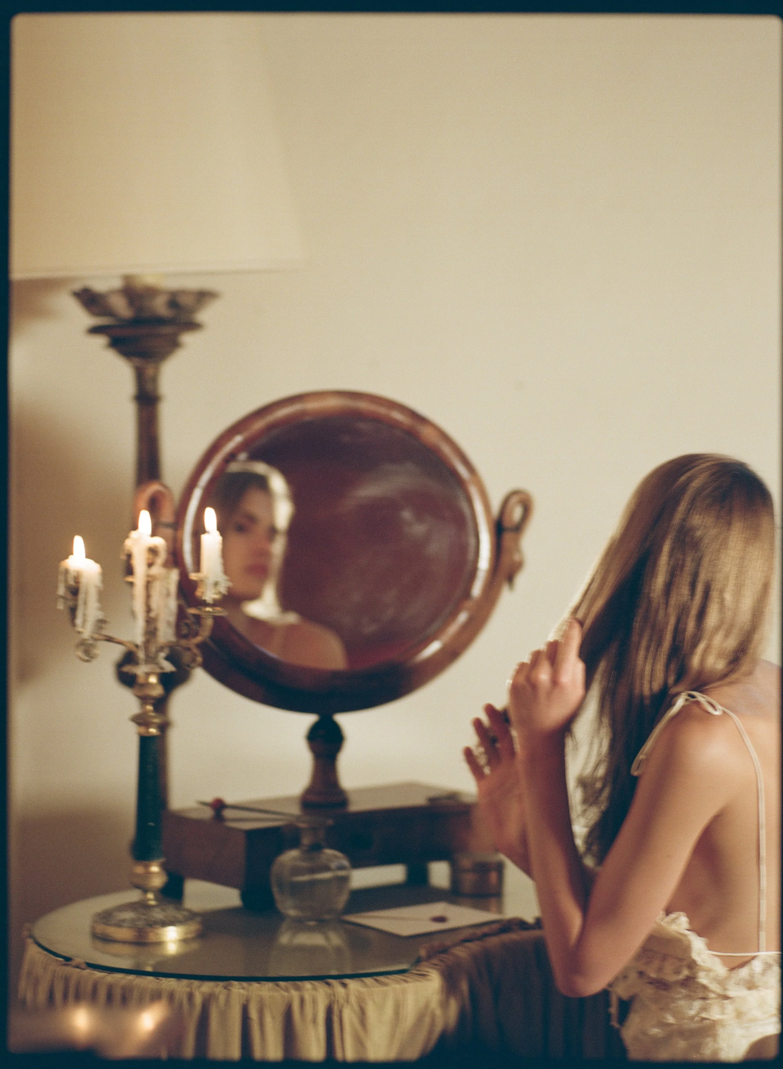 A woman with long blonde hair and wearing a beige dress stands next to a vintage dresser with a mirror, candles, and decorative objects, with her hand touching her hair.