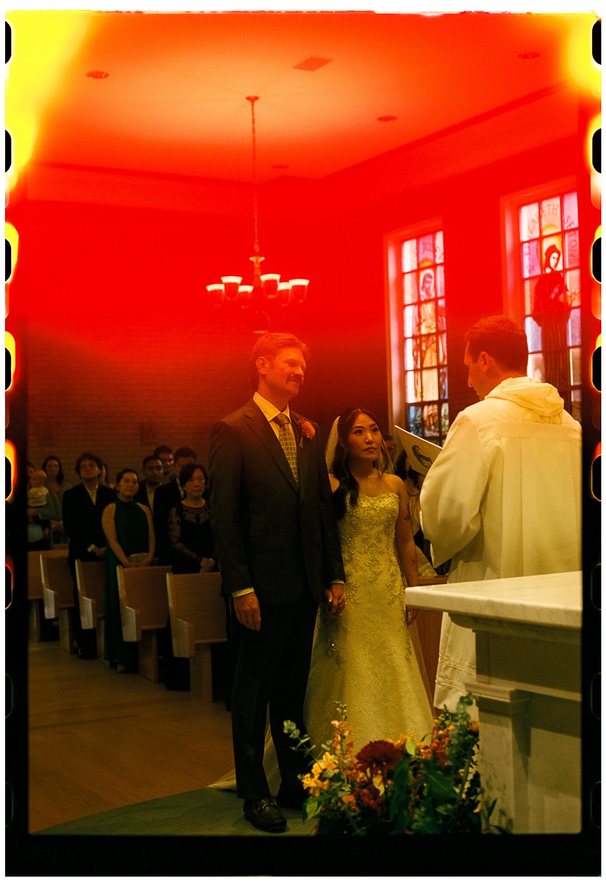 A wedding ceremony inside a church with a bride and groom standing at the altar. The groom is wearing a dark suit, and the bride is in a yellow wedding dress. The priest is wearing white robes. There are stained glass windows depicting saints in the 