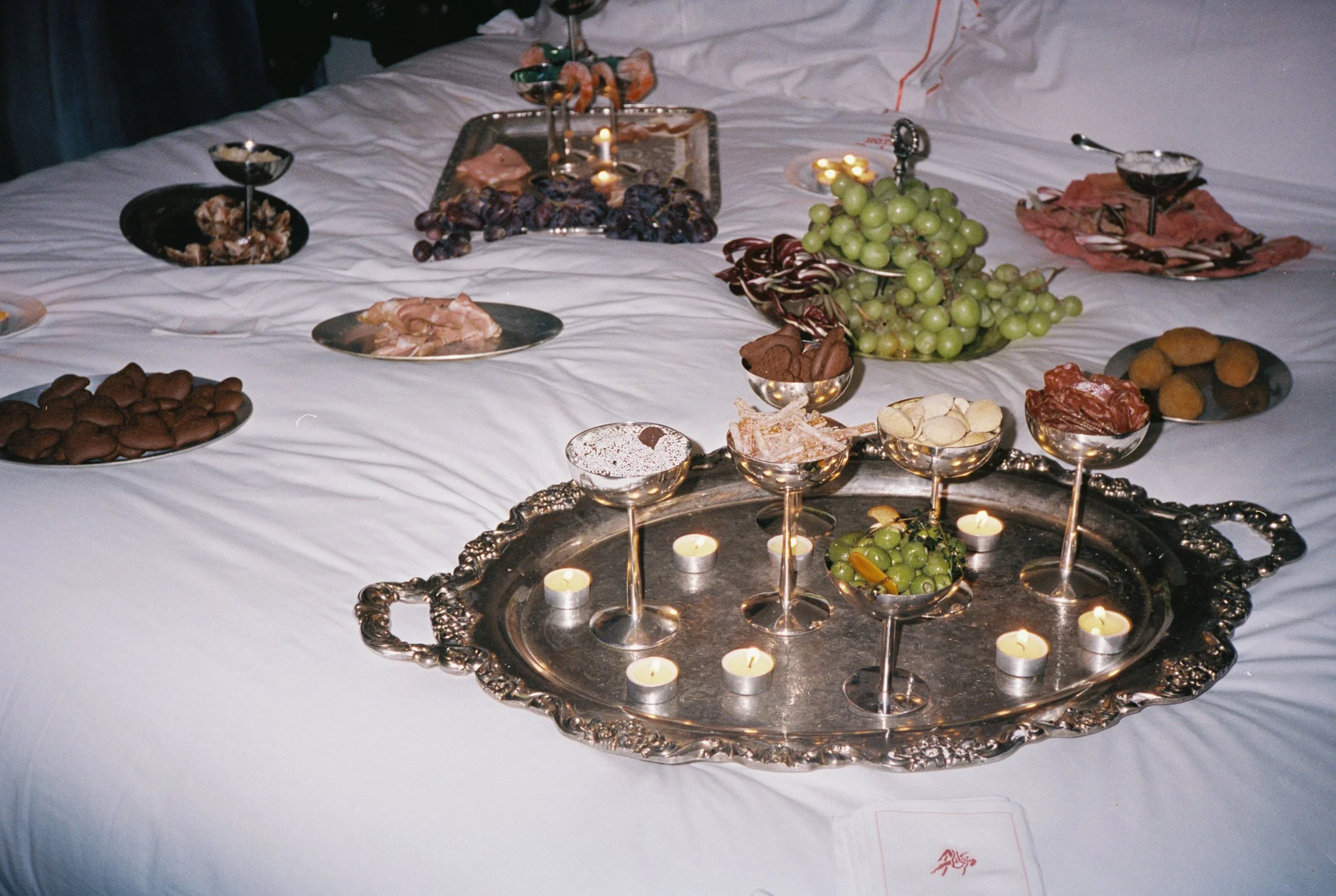 A banquet table with a white tablecloth featuring various fruits, snacks, and candles, including grapes, dates, nuts, and small bowls of candies.