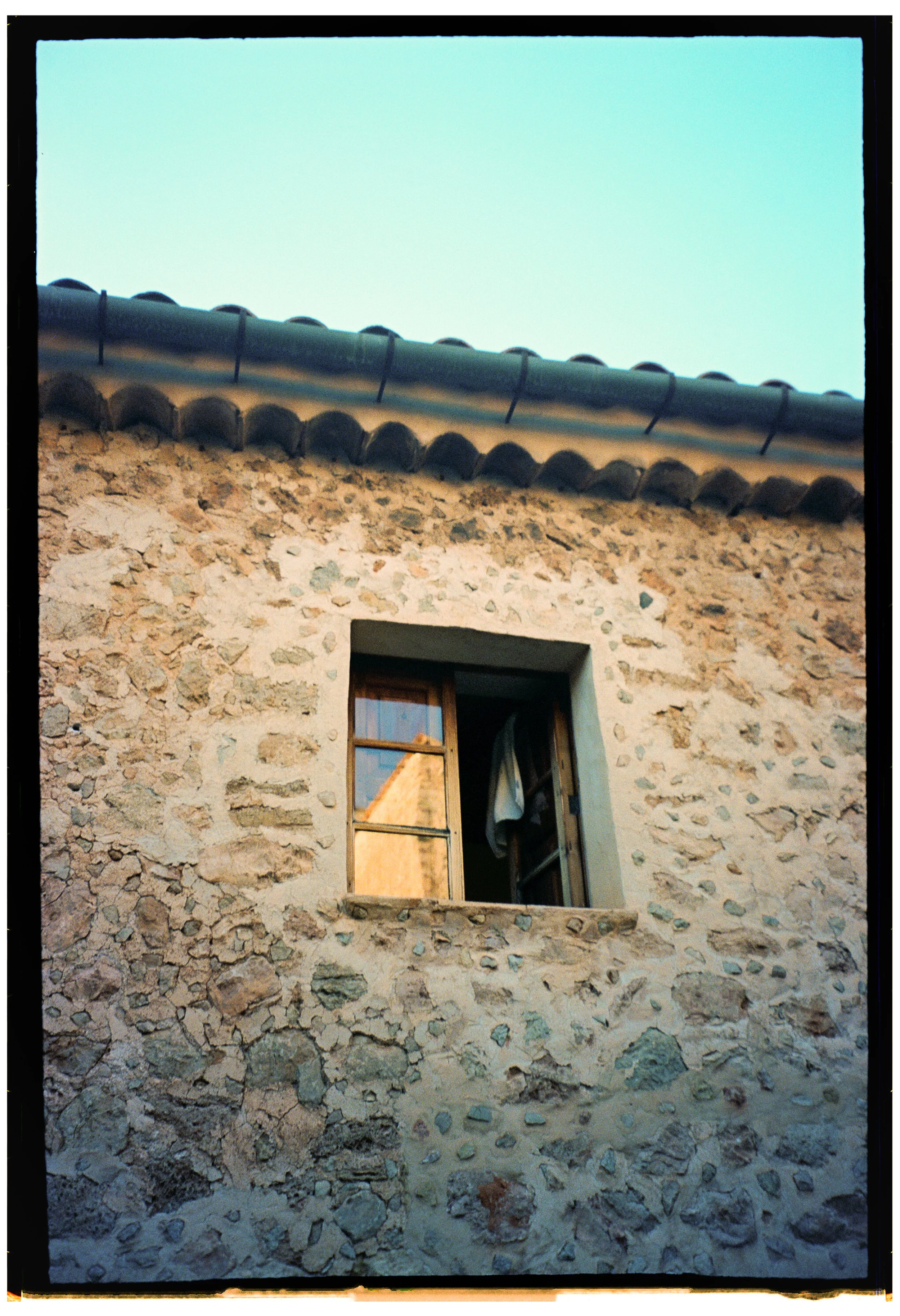 Close-up of a stone building wall with a small window, showing an open window with a curtain inside. The roof has traditional curved tiles, and the sky is visible above.