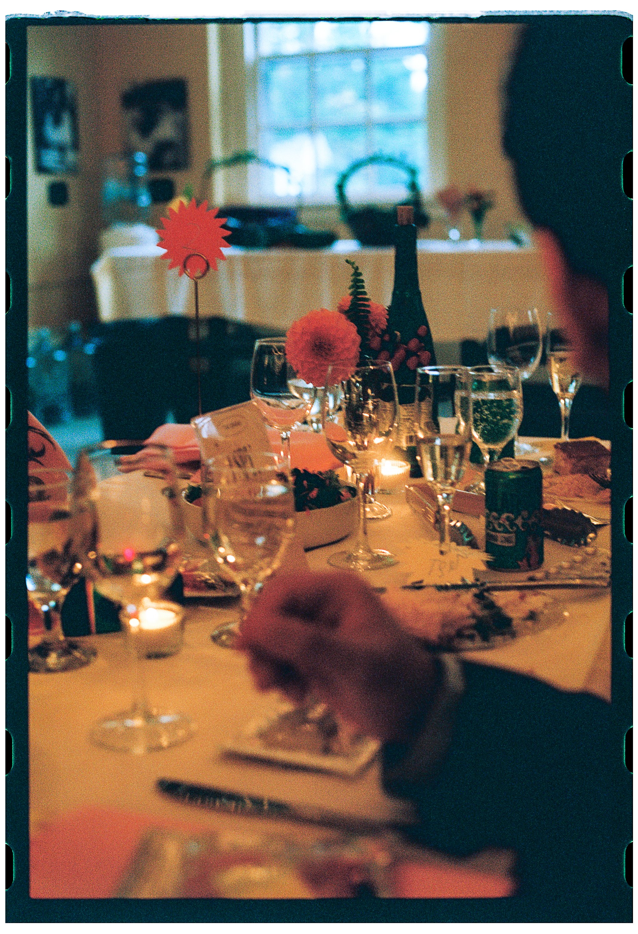 A dining table decorated with flowers, glasses of water, and candles, set for a meal in a cozy room with a window in the background.
