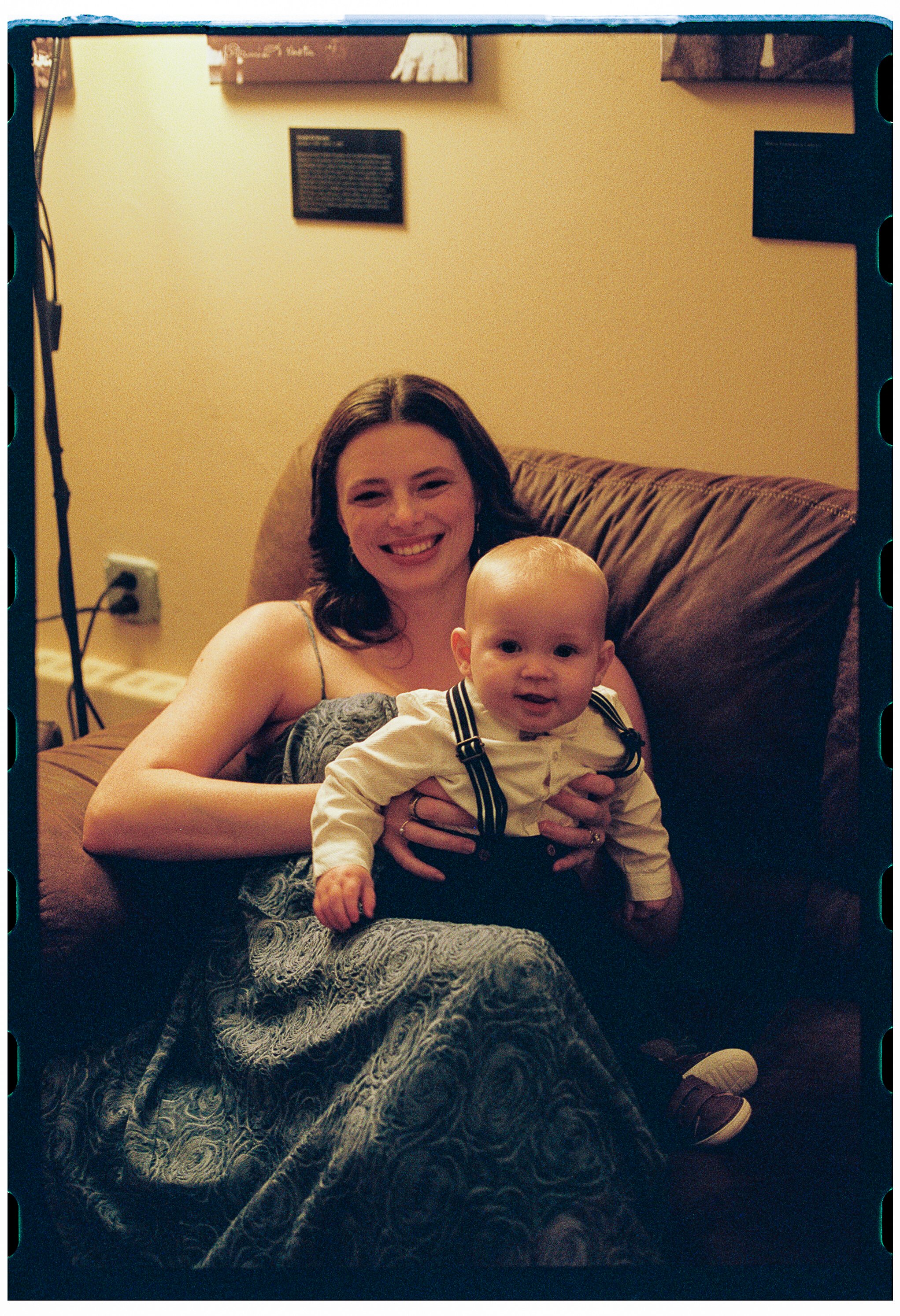 A woman with shoulder-length dark hair smiling while sitting on a brown sofa, holding a young baby boy with light hair. The baby is wearing a cream-colored shirt with black suspenders, and they are in a living room with a beige wall and framed artwor