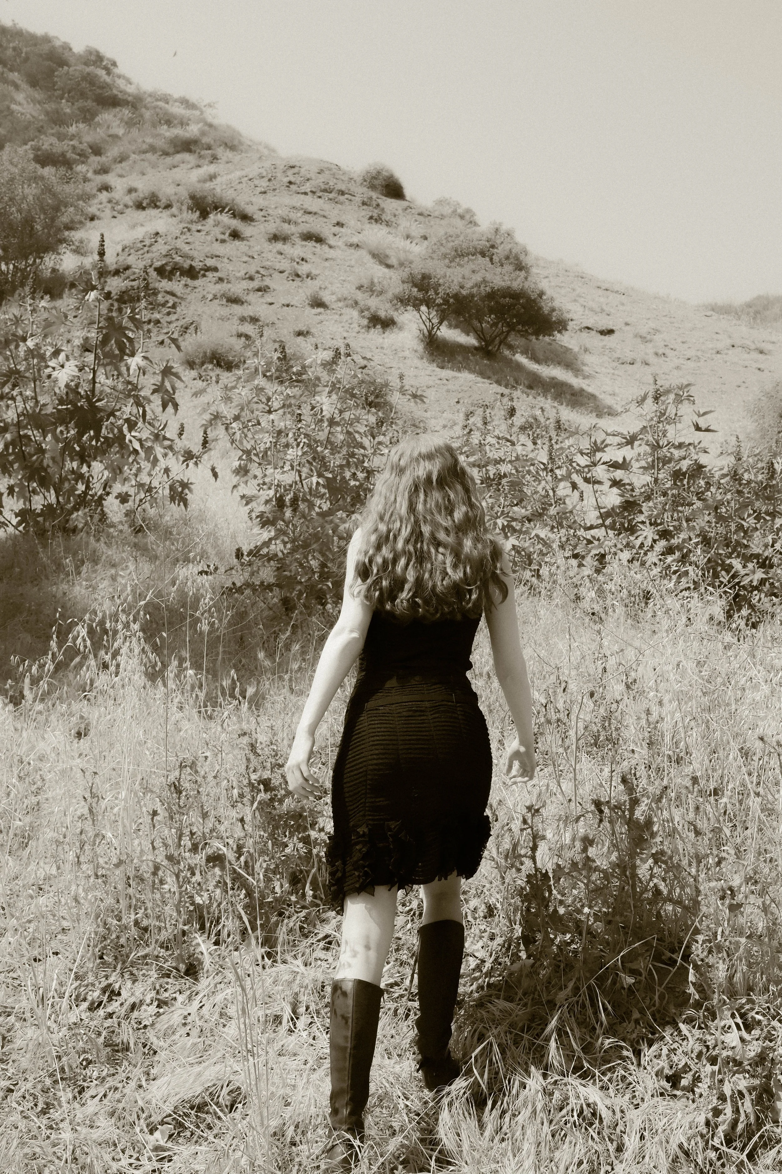 A woman with long wavy hair wearing a black dress and tall black boots walking through a grassy field with bushes and trees on a hillside in the background.