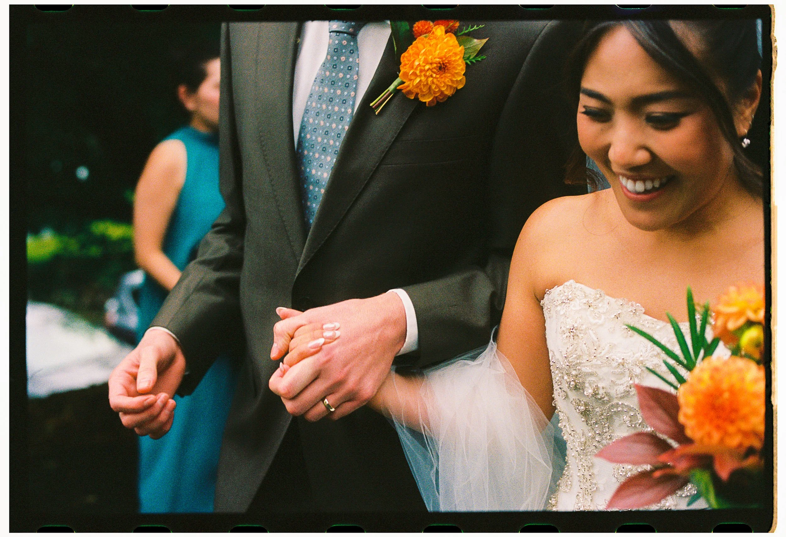 Close-up of a bride and groom holding hands during a wedding ceremony, with the bride smiling and wearing a white lace wedding dress, and the groom in a dark suit with a colorful floral boutonniere.