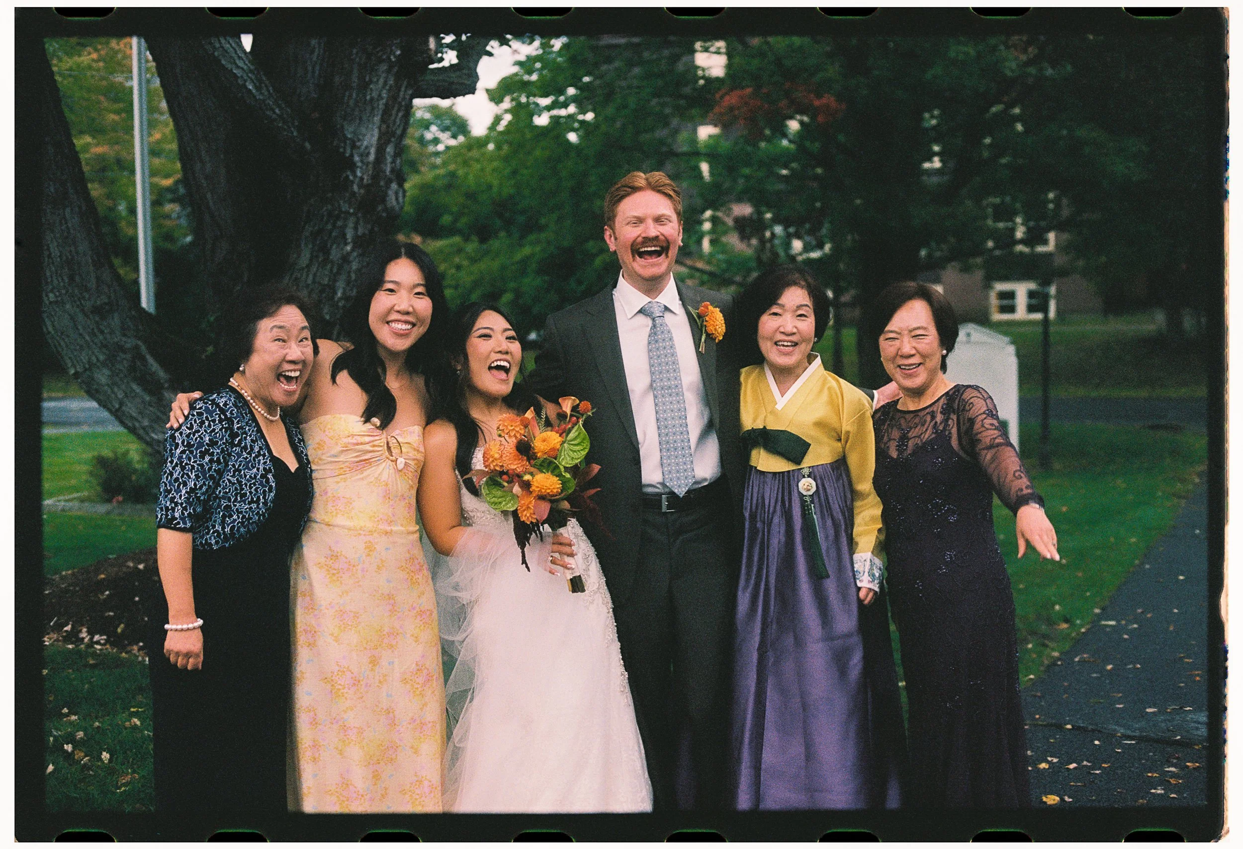 A group of six people, including a woman in a white wedding dress and five women in colorful dresses, standing outdoors by trees during daylight. They are smiling and celebrating a joyful moment.