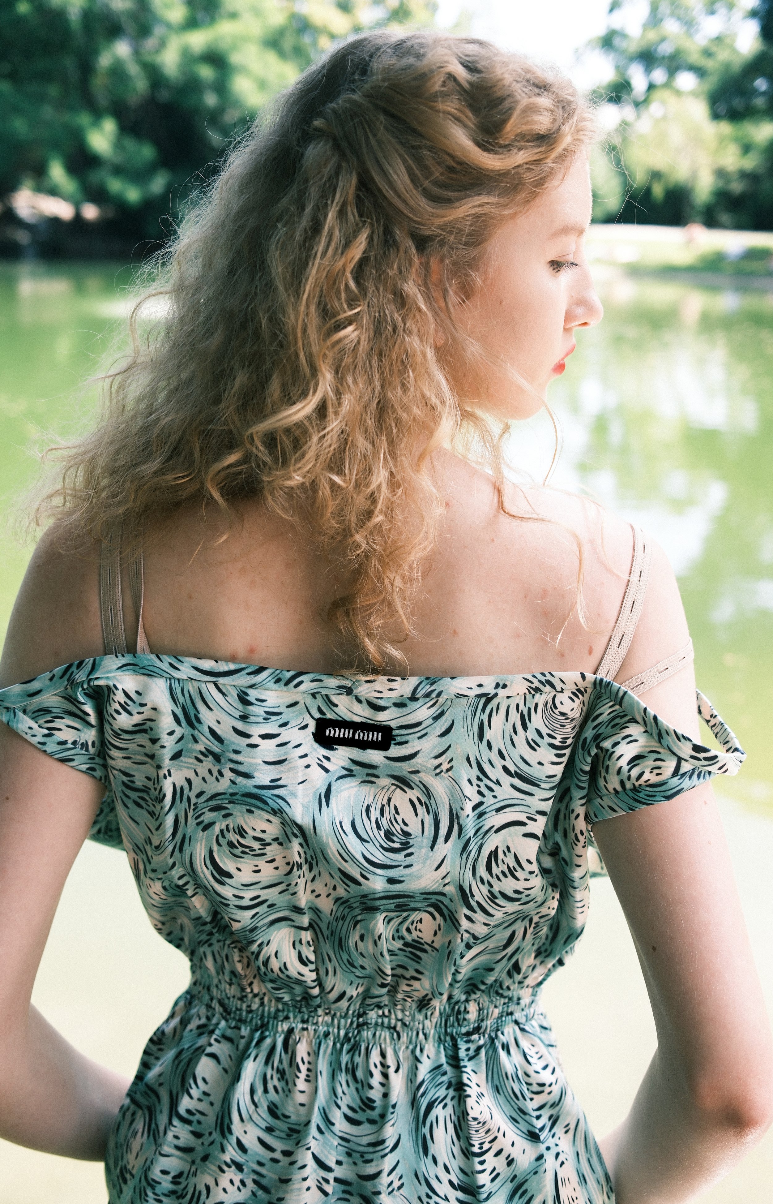 A woman with curly hair standing near a body of water in a park, wearing a patterned dress.