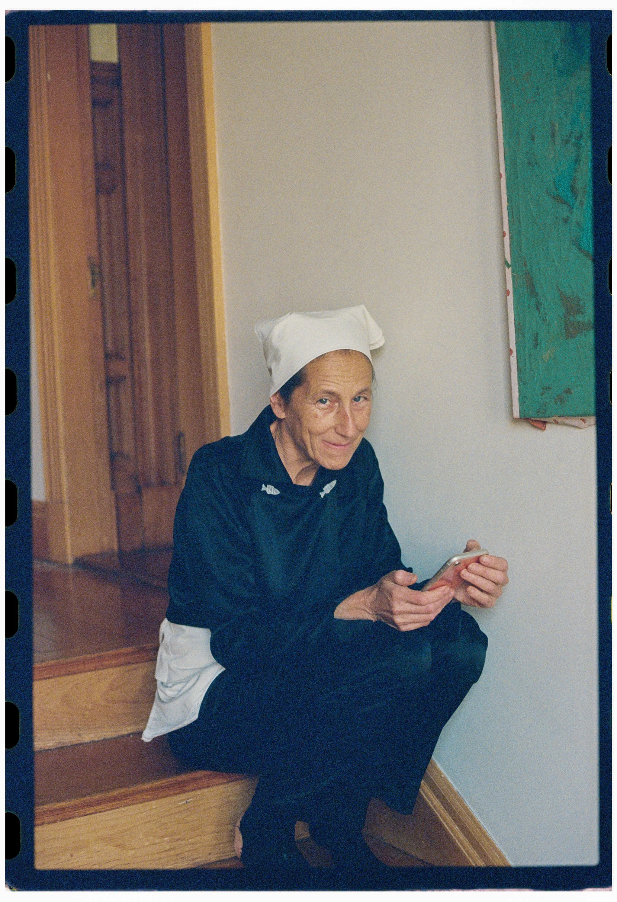 An elderly woman wearing a white nurse's cap and dark clothing is seated on a wooden staircase, smiling while using her phone. The background features a wooden door and a piece of abstract green artwork on the wall.