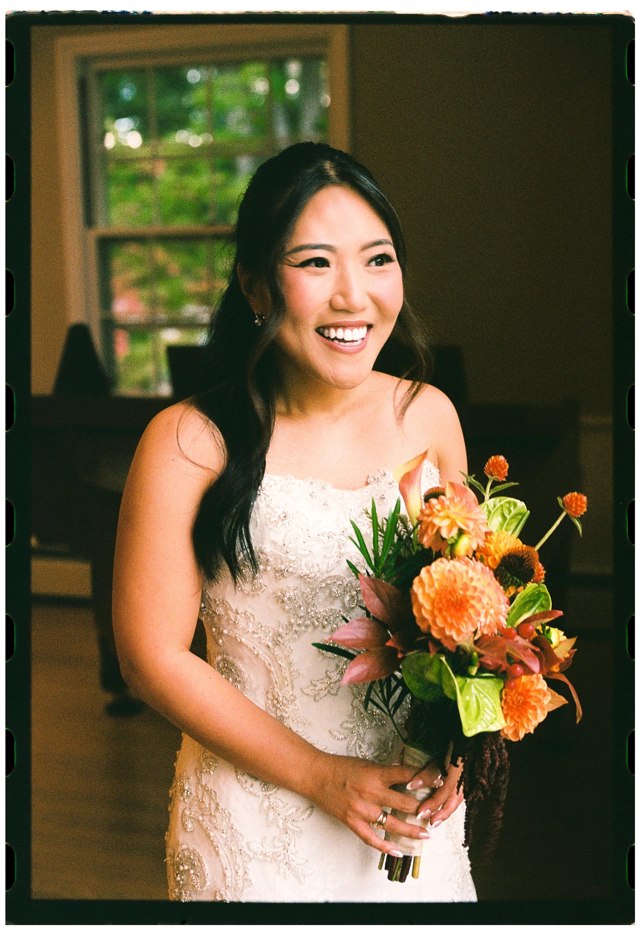 A smiling woman in a wedding dress holding a bouquet of orange and green flowers in a room with a window showing trees outside.