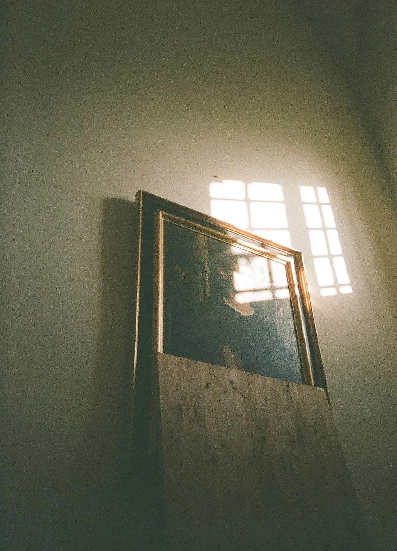 A framed picture partly visible on a wall, with sunlight coming through a window, creating a grid of light and shadow.