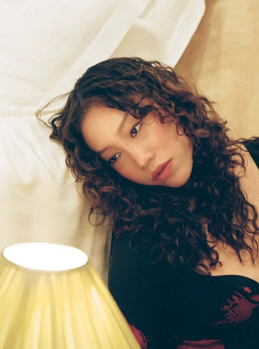 A woman with curly hair, wearing a black top, reclining against a white headboard in a softly lit room.
