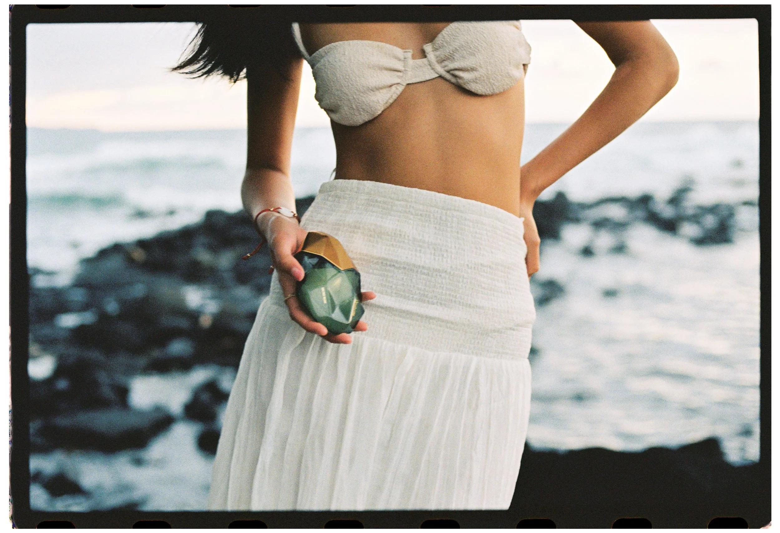 A woman wearing a beige, textured top and a high-waisted beige skirt standing near the water, holding a seashell in her right hand at the beach.