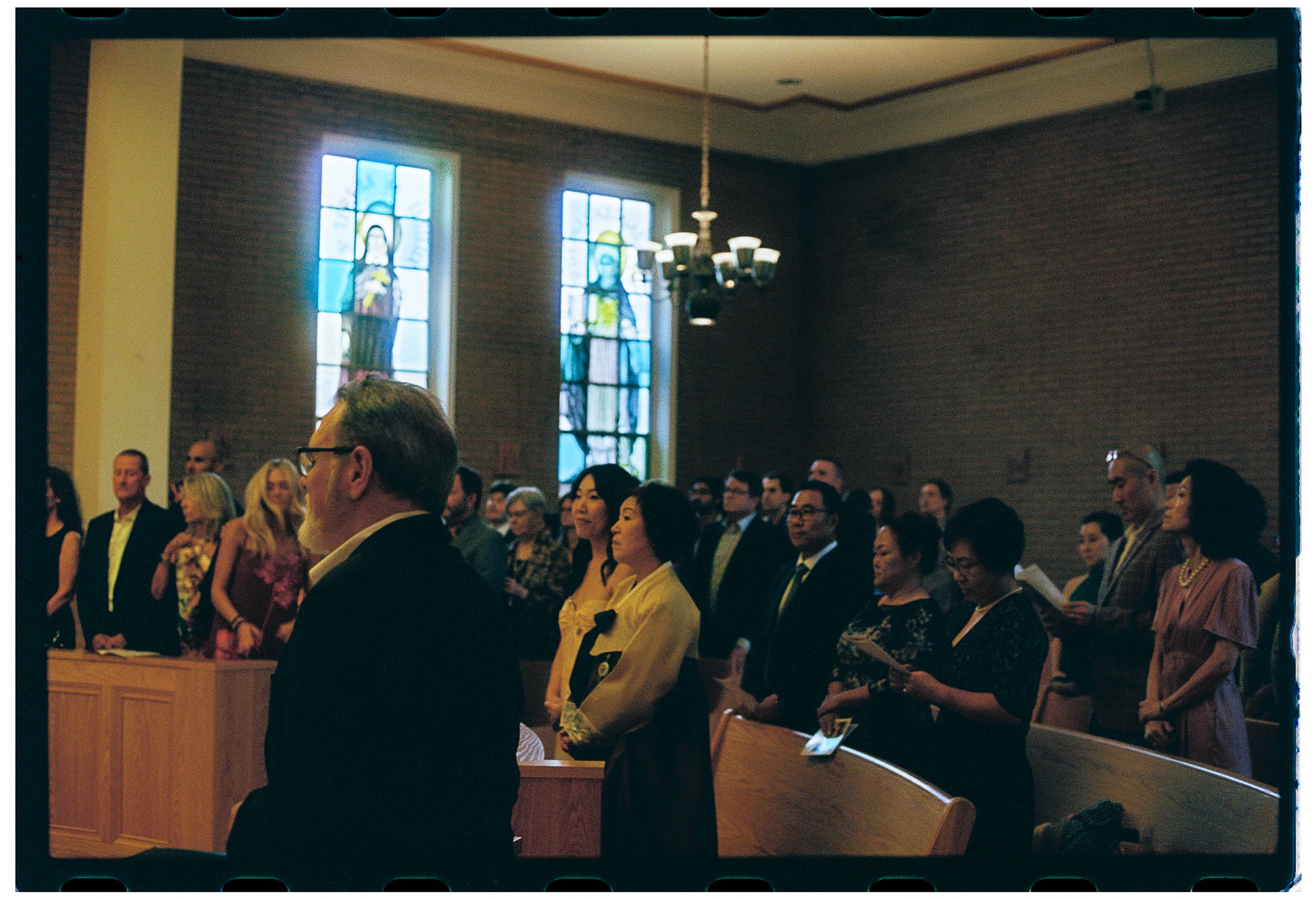 People gathered inside a church during a religious service, with stained glass windows depicting religious figures on the wall behind them.