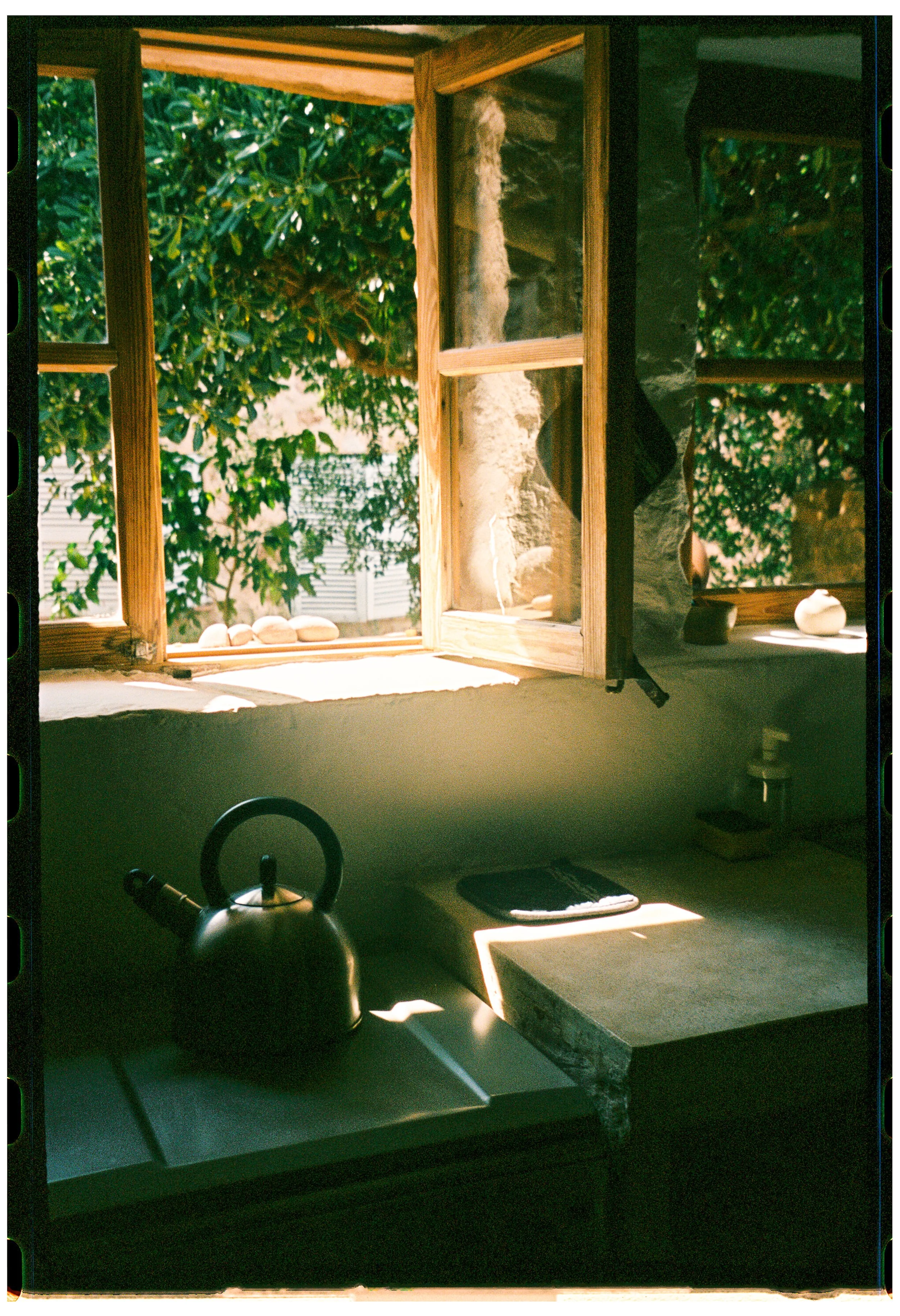 A kitchen windowsill with several round stones, a black kettle, a cutting board, and sunlight streaming through an open window onto greenery outside