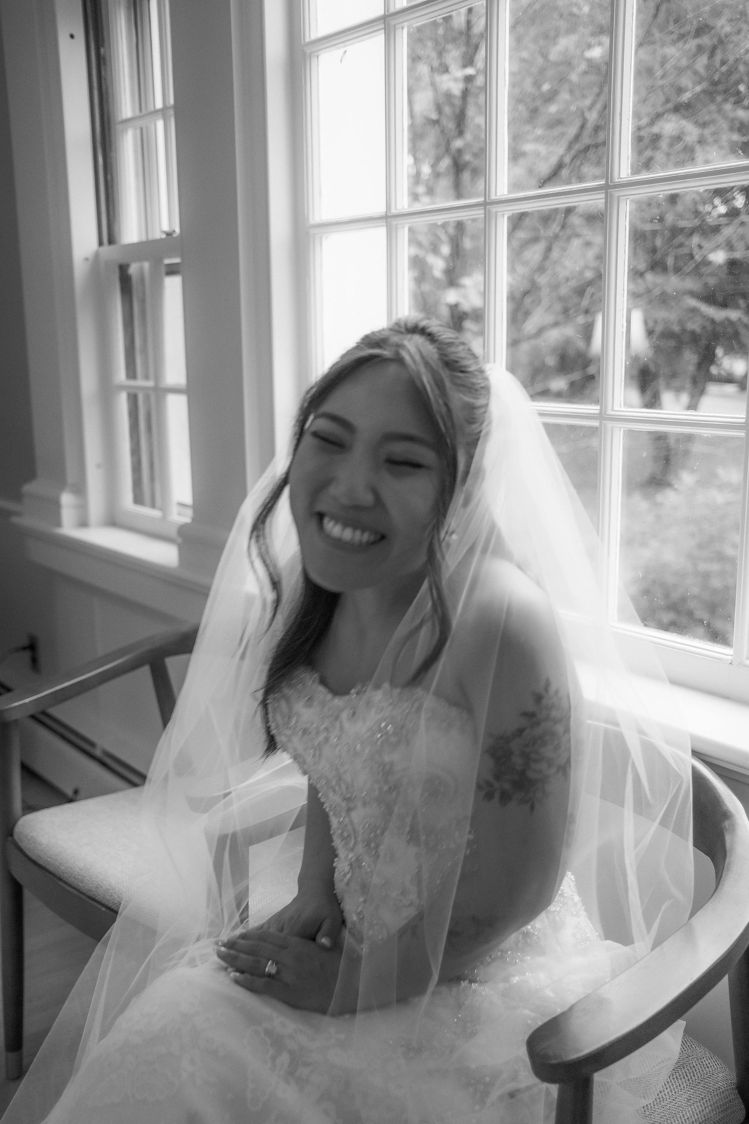A woman dressed in a wedding gown, sitting on a chair near a window with a smile on her face. She is wearing a veil, and the background shows trees outside.