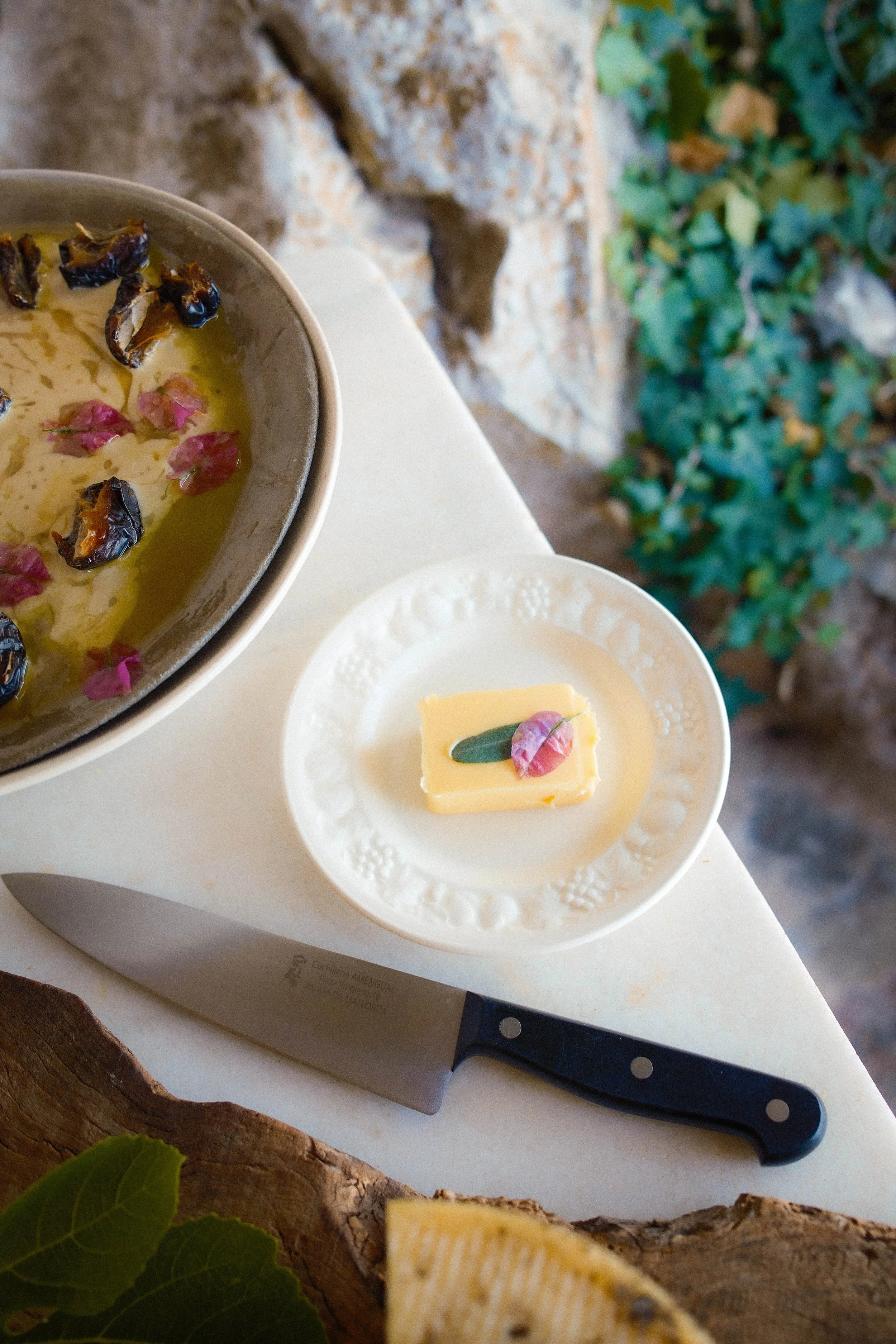A small dish garnished with a bay leaf and pink flowers, a sharp kitchen knife with a black handle on a wooden cutting board, and a bowl of olive oil with herbs and rose petals, set on a white surface with a stone wall and green ivy in the background