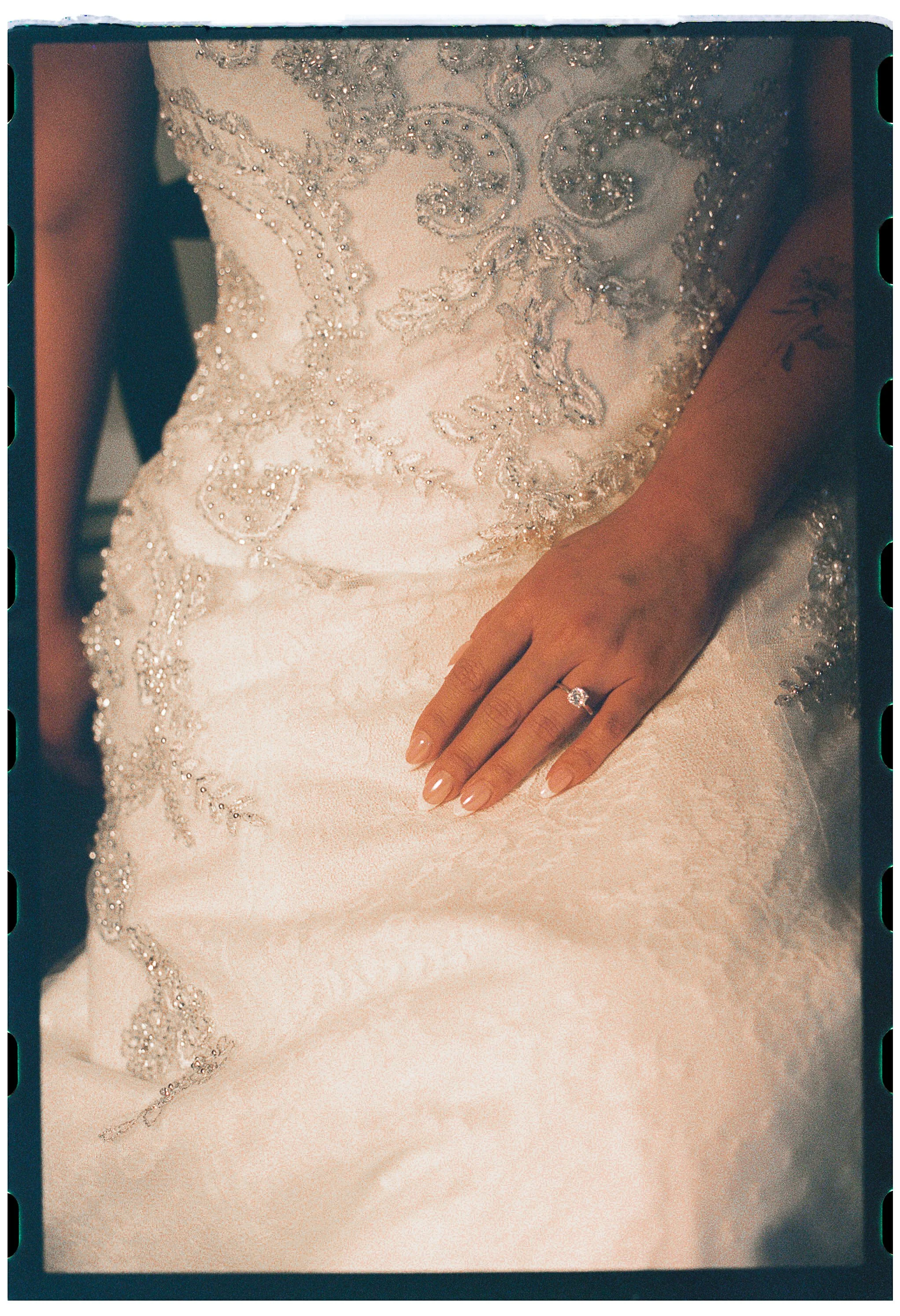 Close-up of a bride's torso wearing a wedding dress with intricate beading, and a hand with a wedding ring resting on the fabric.