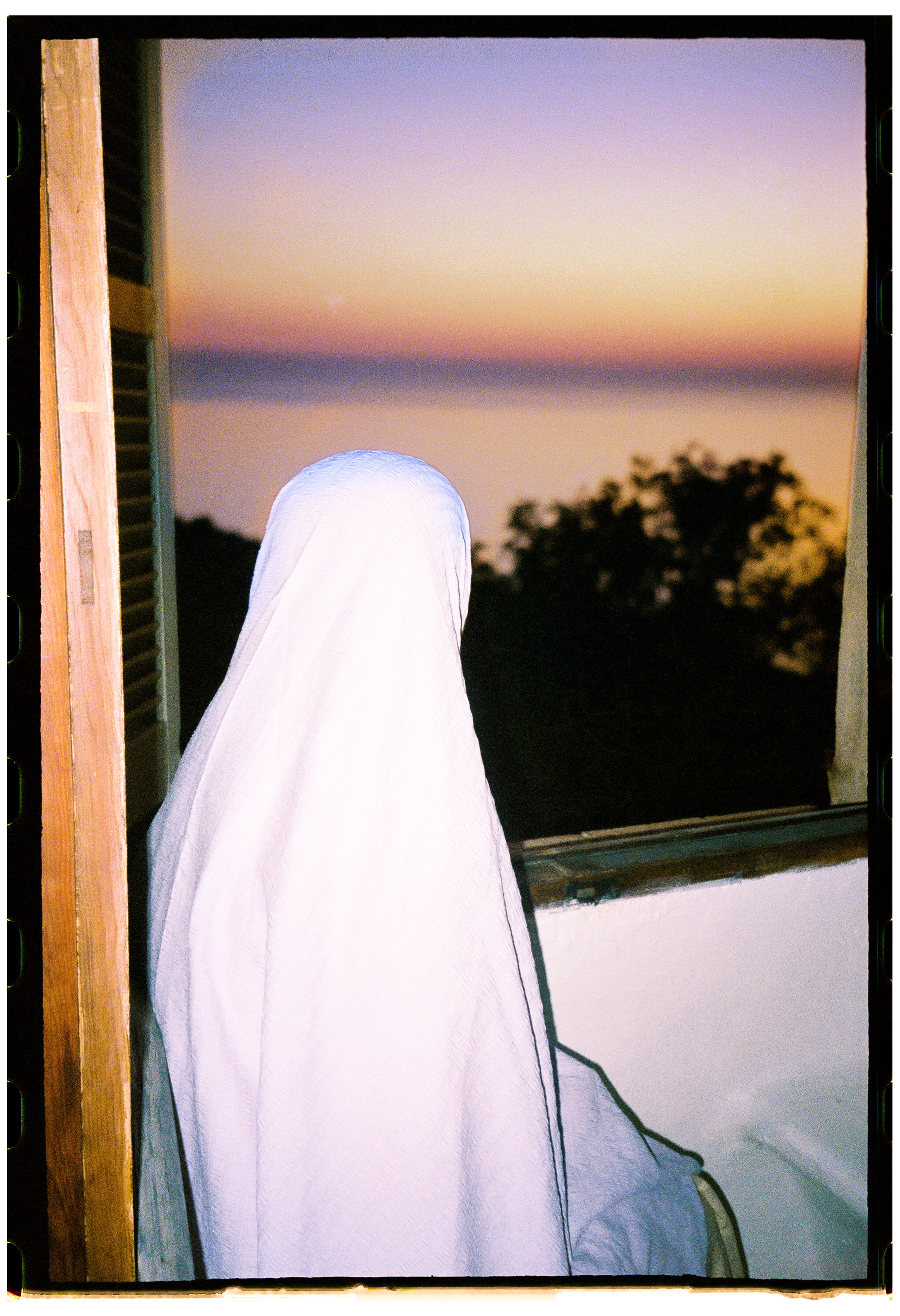 A person dressed in white sitting near an open window during sunset, with a view of trees outside.