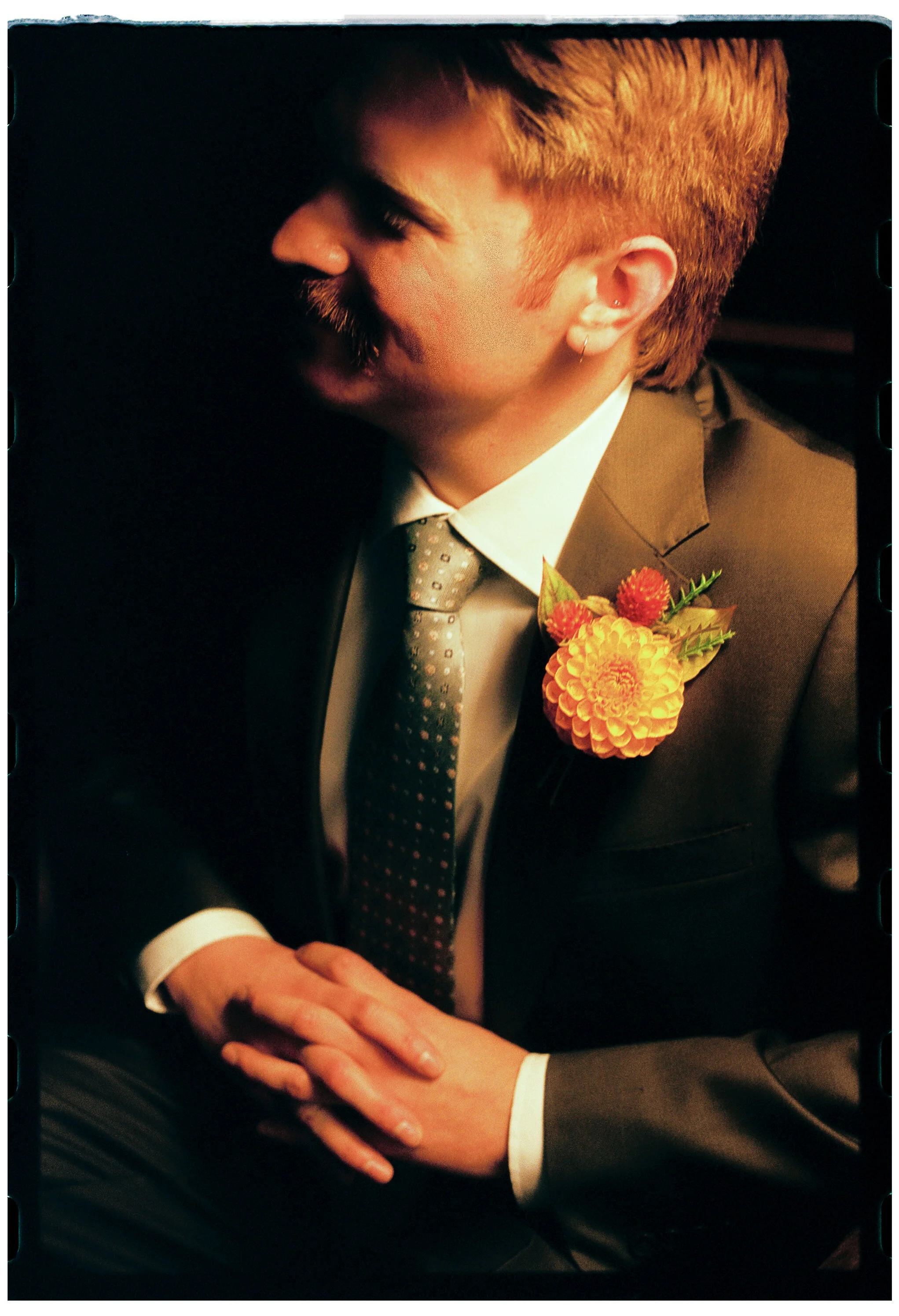 A man with red hair and a beard wearing a grey suit, white shirt, and patterned tie, with a yellow and orange floral boutonniere on his suit lapel, holding hands with someone.