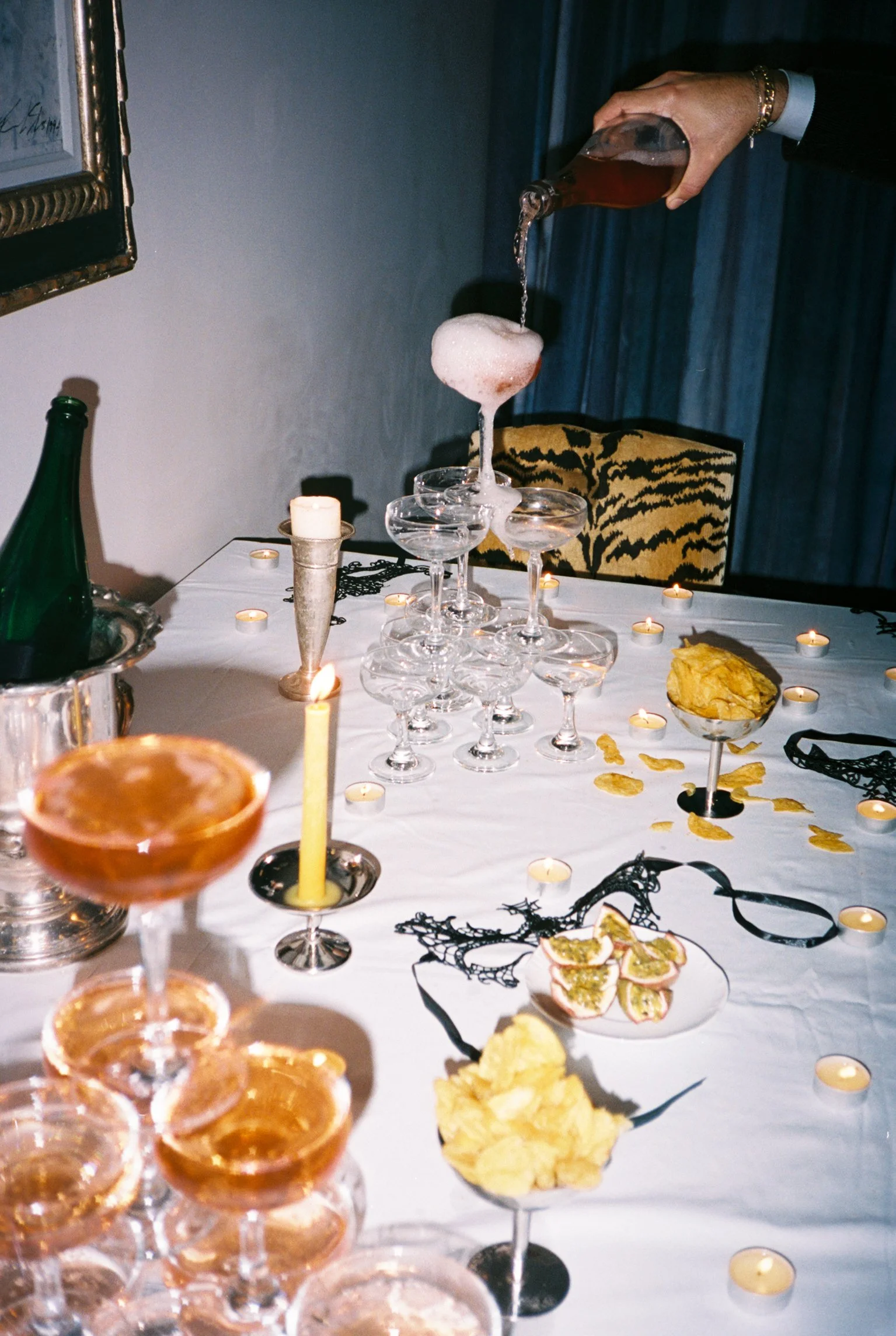 A person pouring a drink into a tower of glasses at a decorated party table with candles, snacks, and black decorative elements.