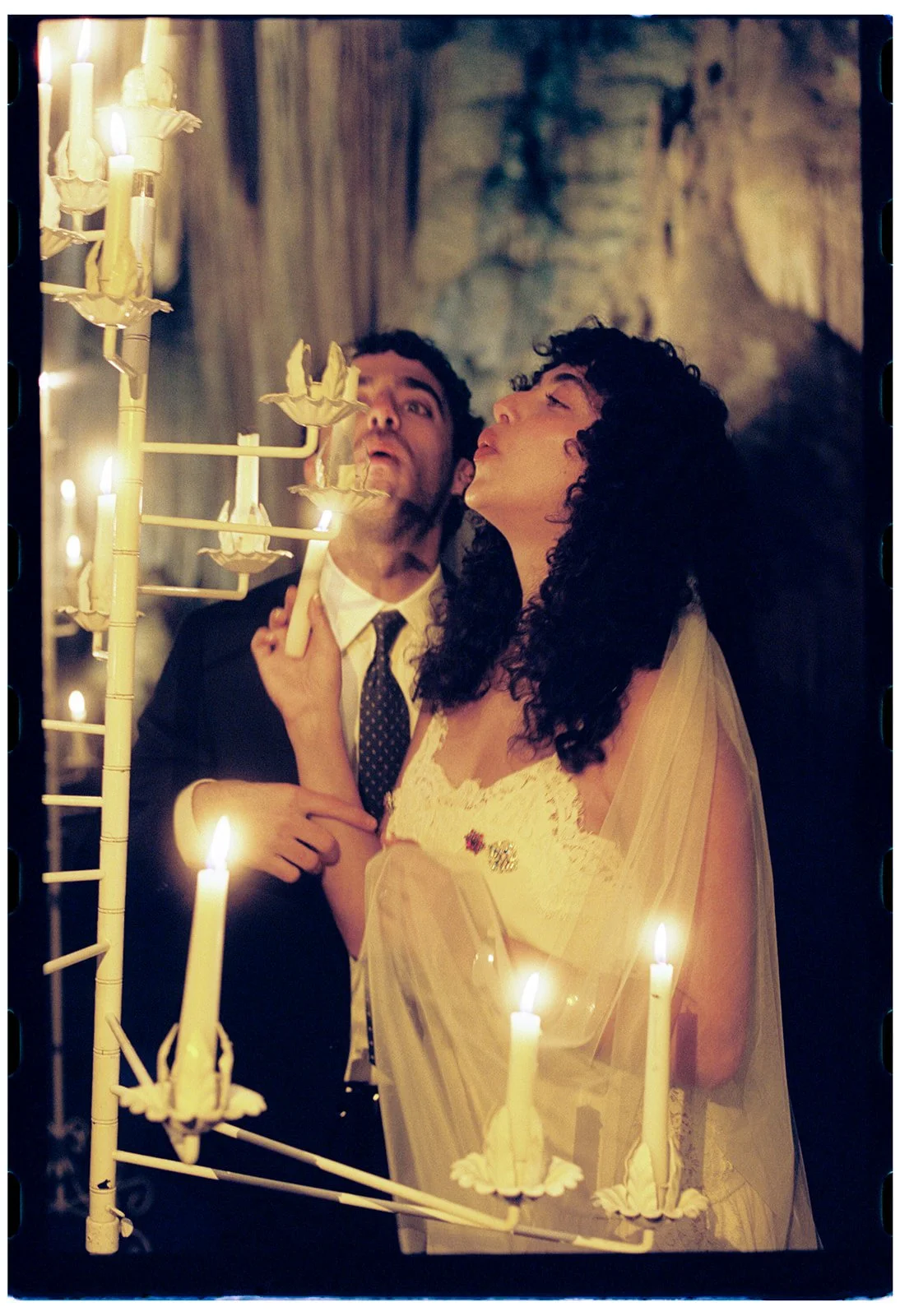 A man and woman dressed in formal attire, standing close together in dim lighting, with candles illuminating their faces, in what appears to be a cave or underground setting.