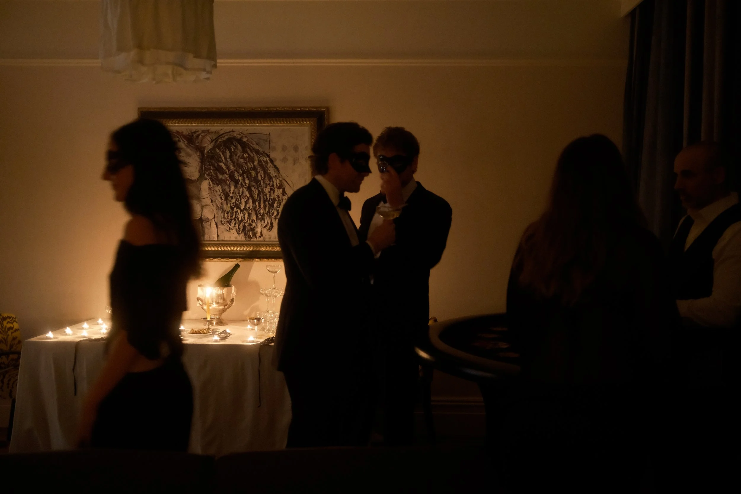 People at a dinner party wearing masks, standing near a table with drinks and candles, in a dimly lit room with a painting on the wall.