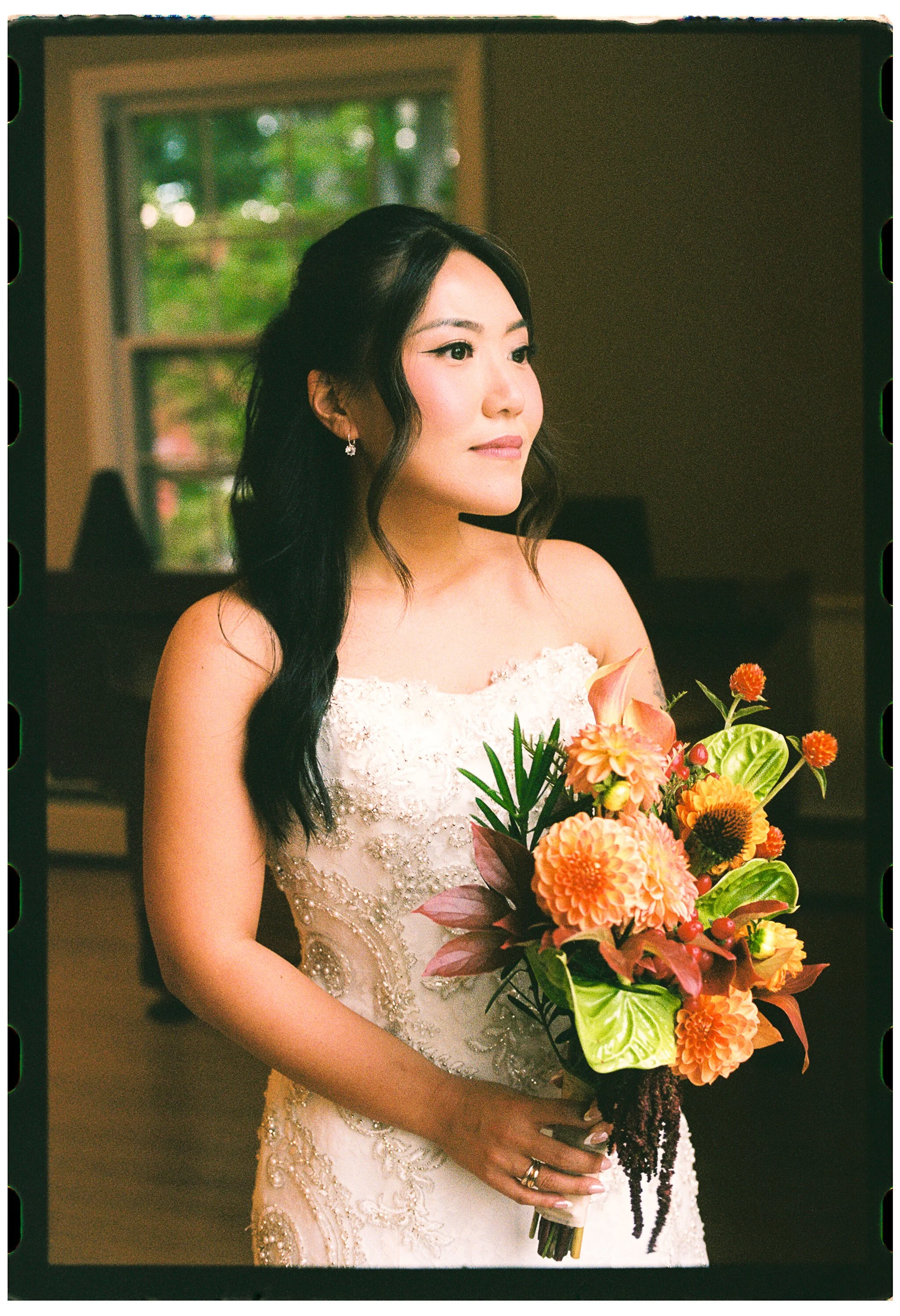 A woman in a white, ornate wedding dress holding a bouquet of colorful flowers, posing indoors near a window with green trees outside.