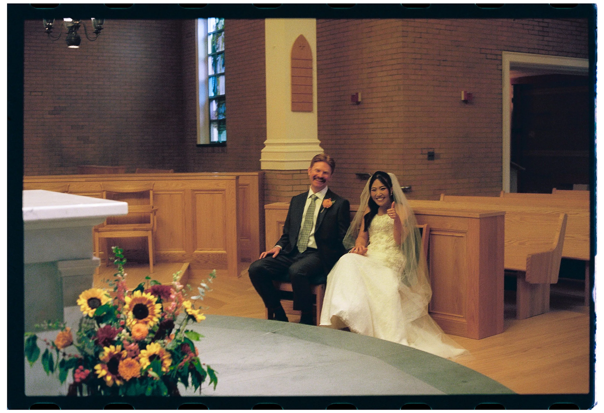 A couple dressed in wedding attire sitting on a wooden pew inside a church, smiling, with a bouquet of sunflowers and flowers on a table in the foreground.