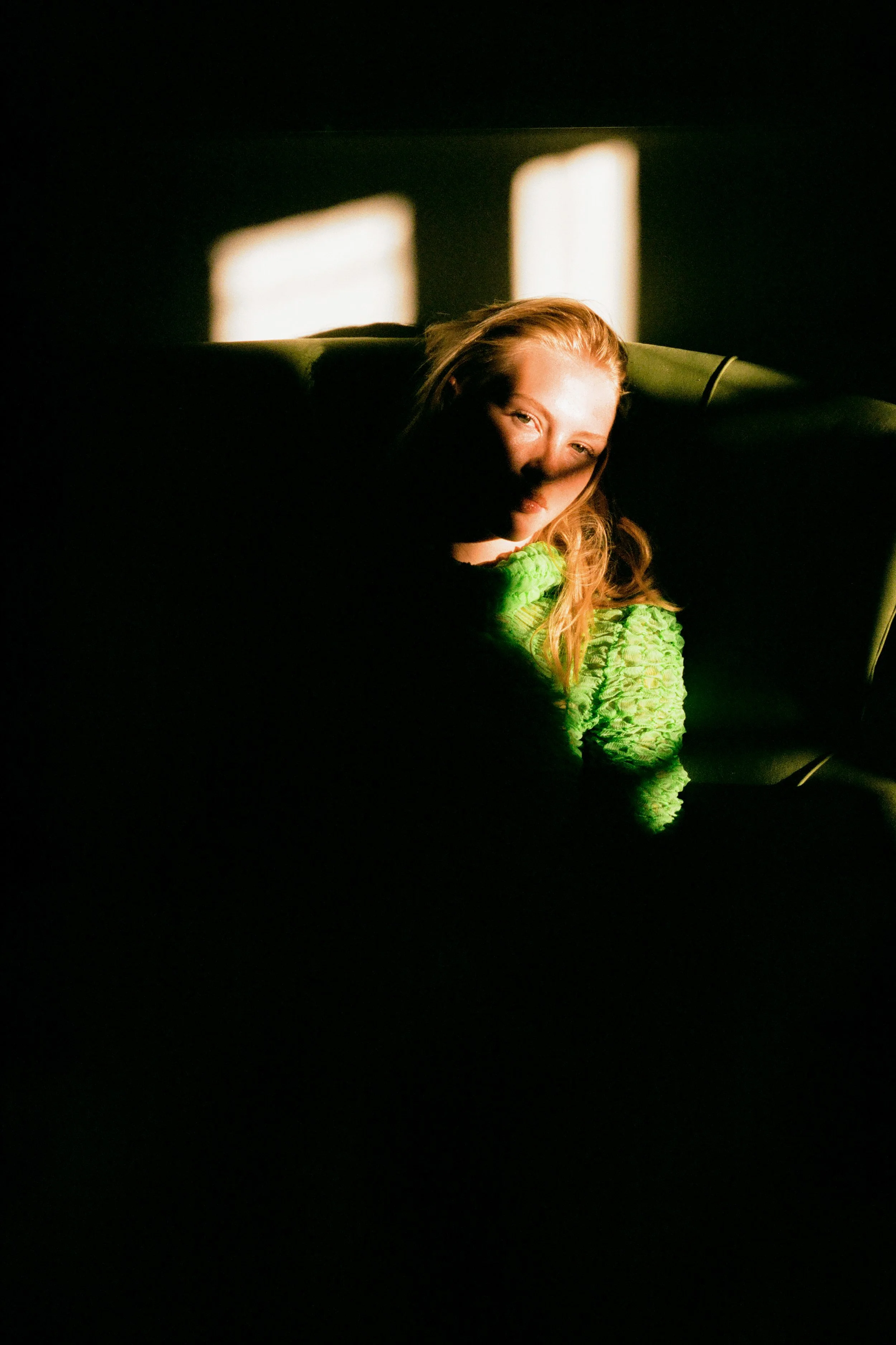 A woman with red hair wearing a bright green top, resting her head on a dark surface, illuminated by a soft, warm light with blurred windows or light sources in the background.