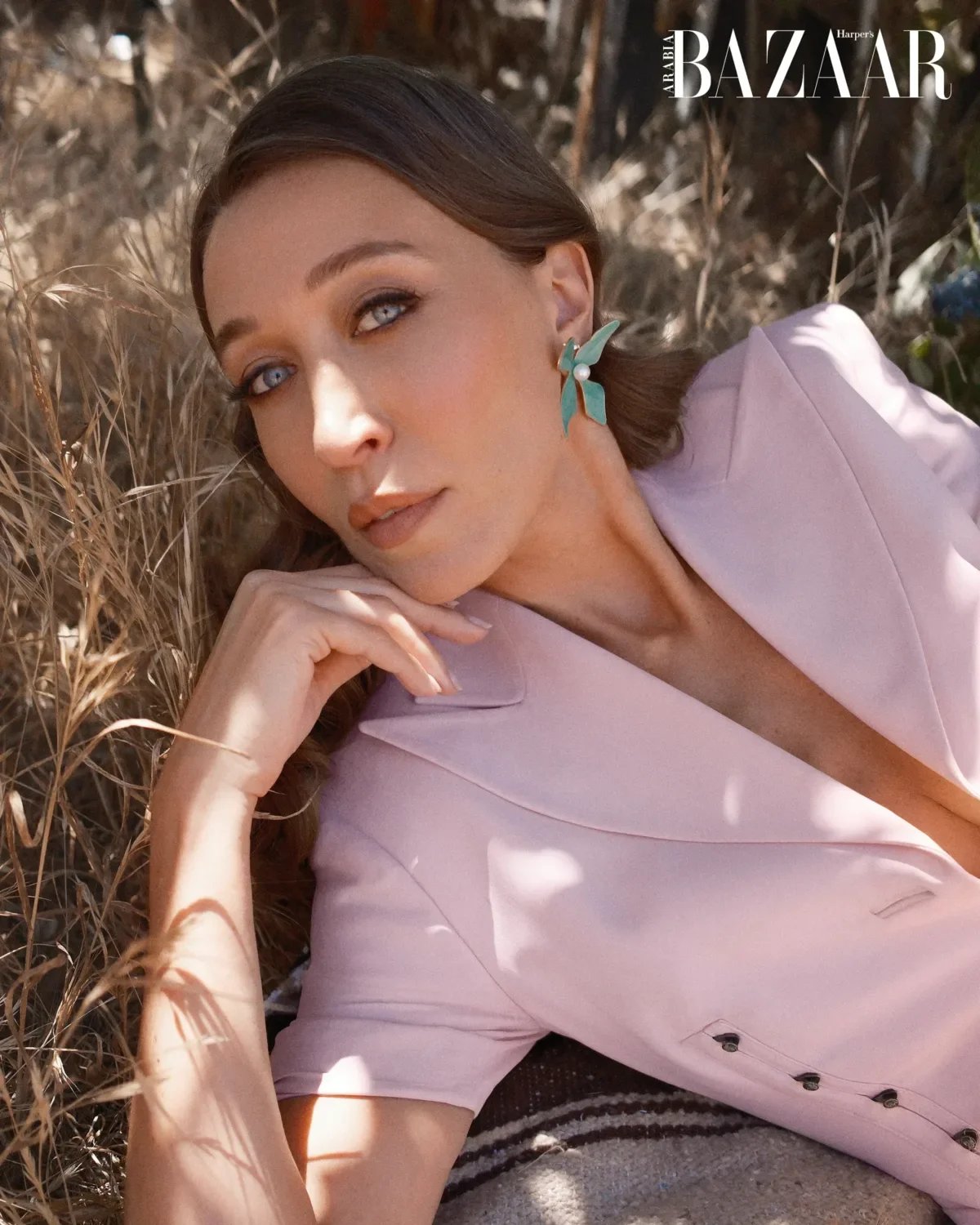 A woman with blue eyes, wearing turquoise earrings with a pearl, lying on the ground surrounded by dried grass. She has brown hair and is dressed in a light pink blazer, resting her chin on her hand.