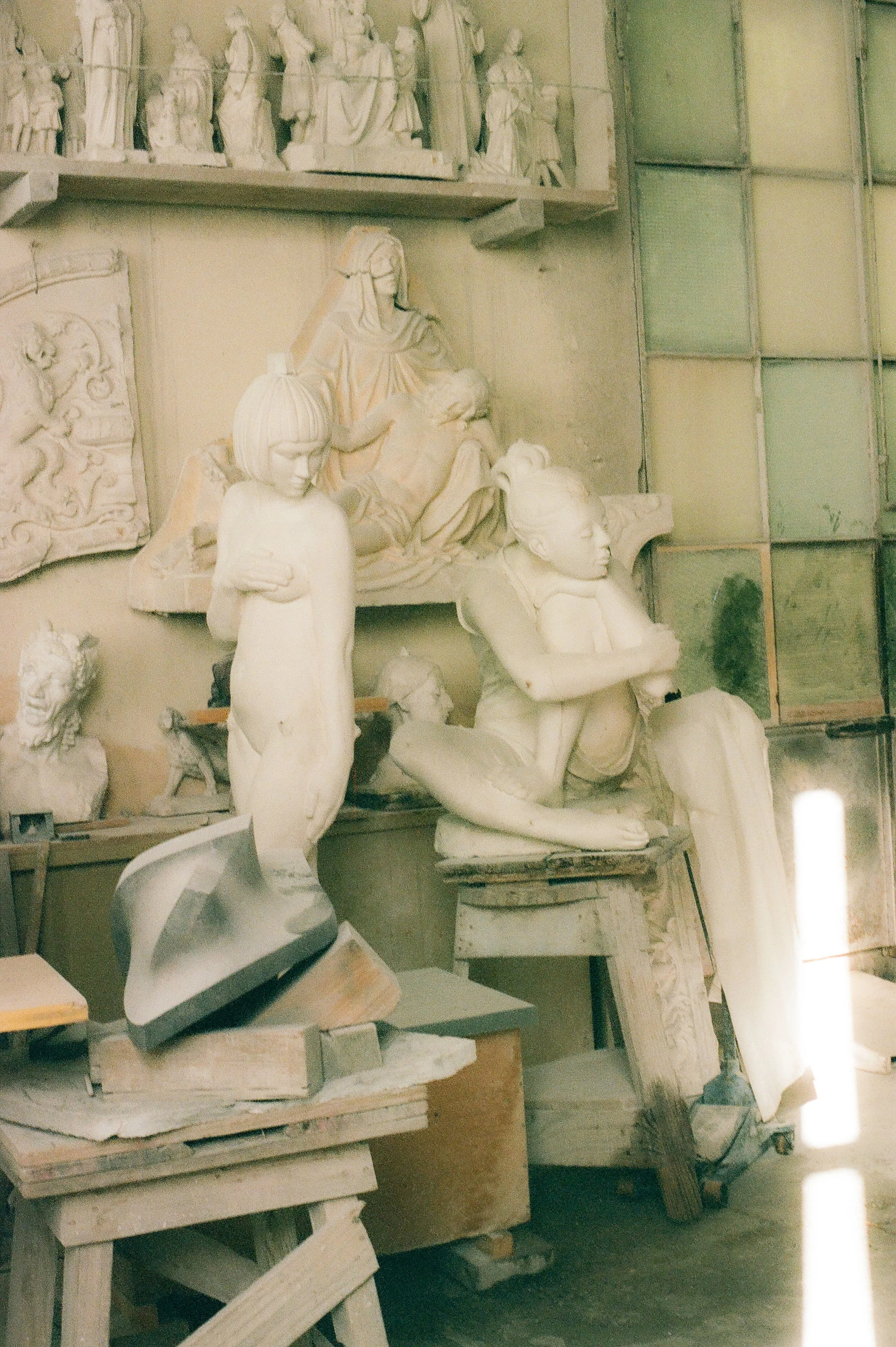 A workshop with various unfinished sculptures and relief carvings, including figures and faces, surrounded by tools and wooden supports.