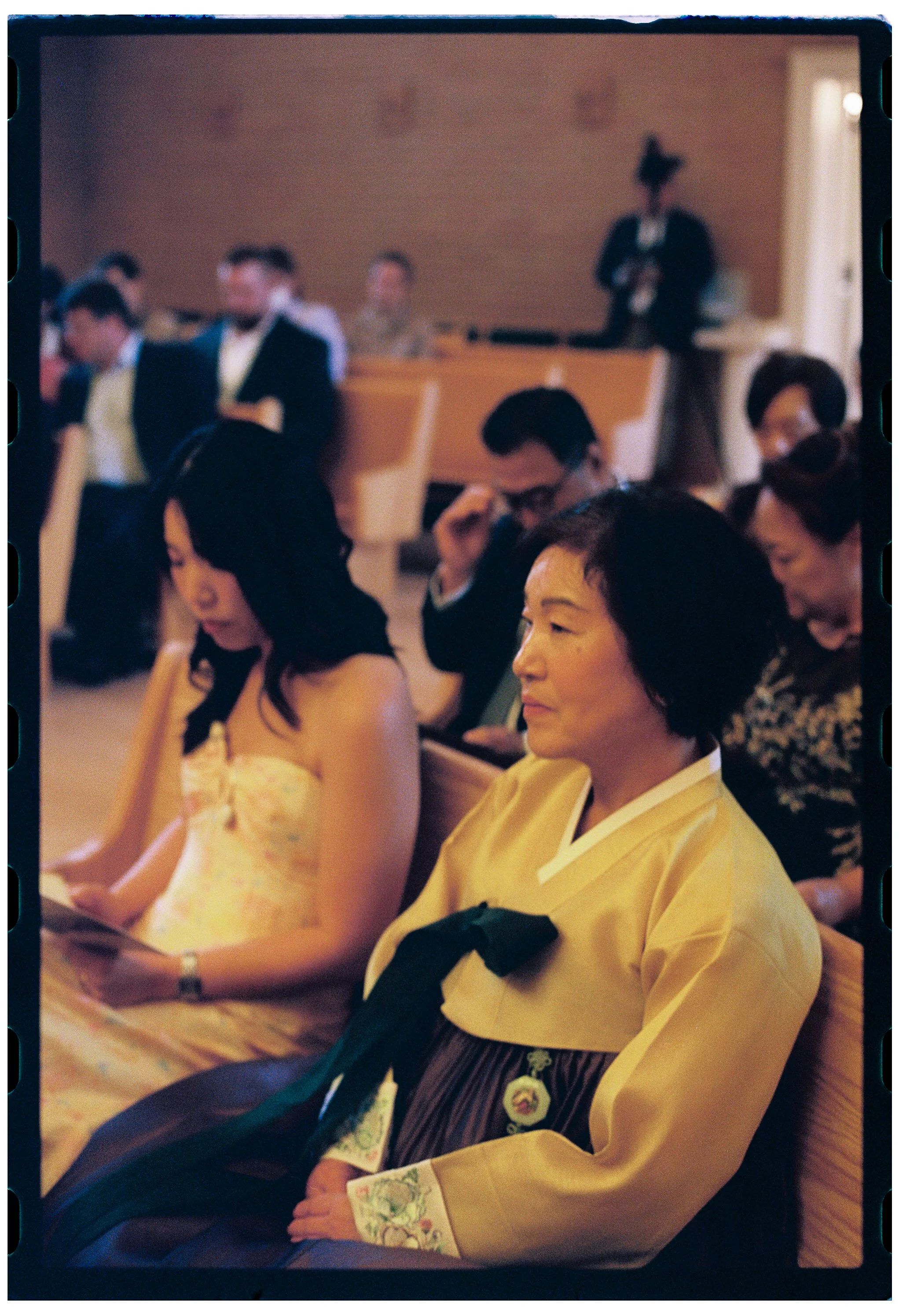 A group of people attending a formal event, seated in a spacious room with warm lighting. The woman in the foreground wears traditional Japanese attire, including a yellow kimono with floral accents, and appears to be focused, possibly watching a per