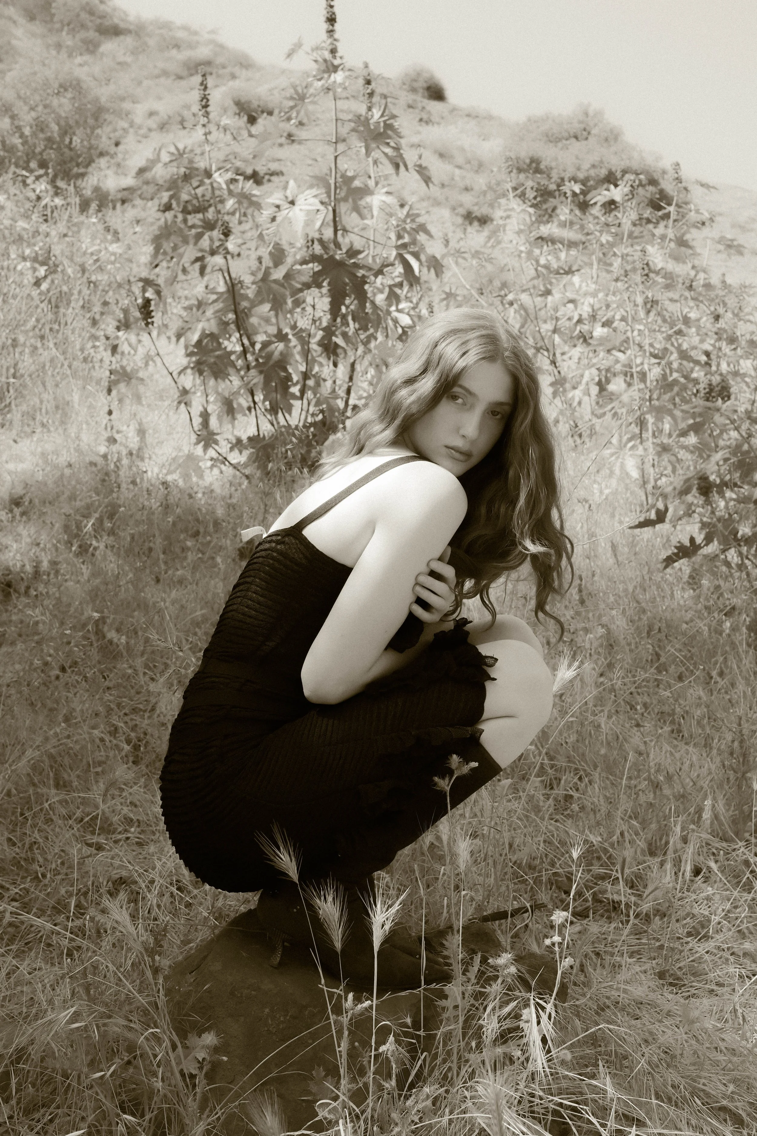 A young woman with long, wavy hair kneeling in a grassy field with flowers, looking at the camera. Background features grassy hills and bushes, black and white photo.