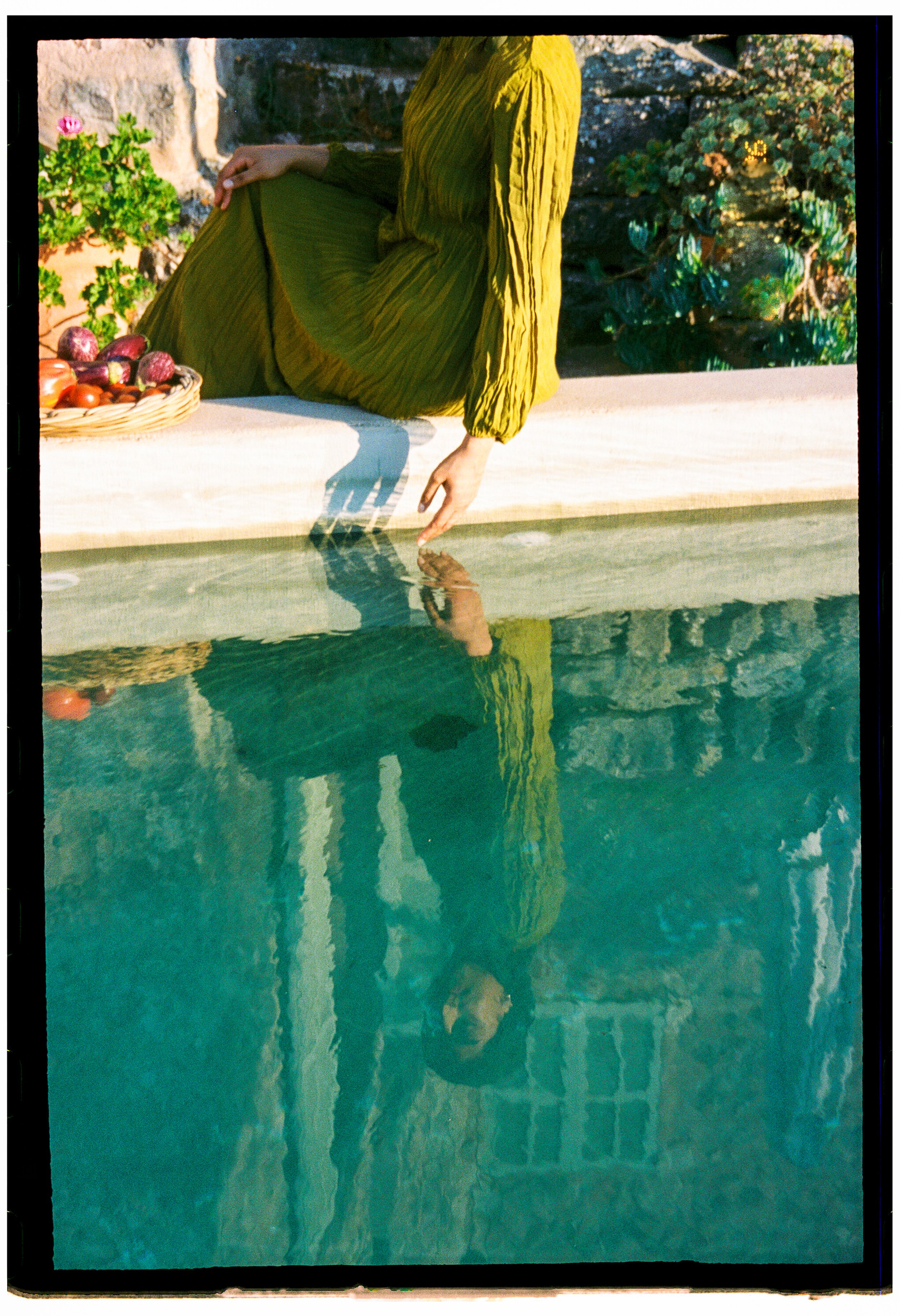 Person wearing a green dress sitting on a ledge by a swimming pool, reaching into the water with their hand, with a basket of fruits nearby and greenery in the background.