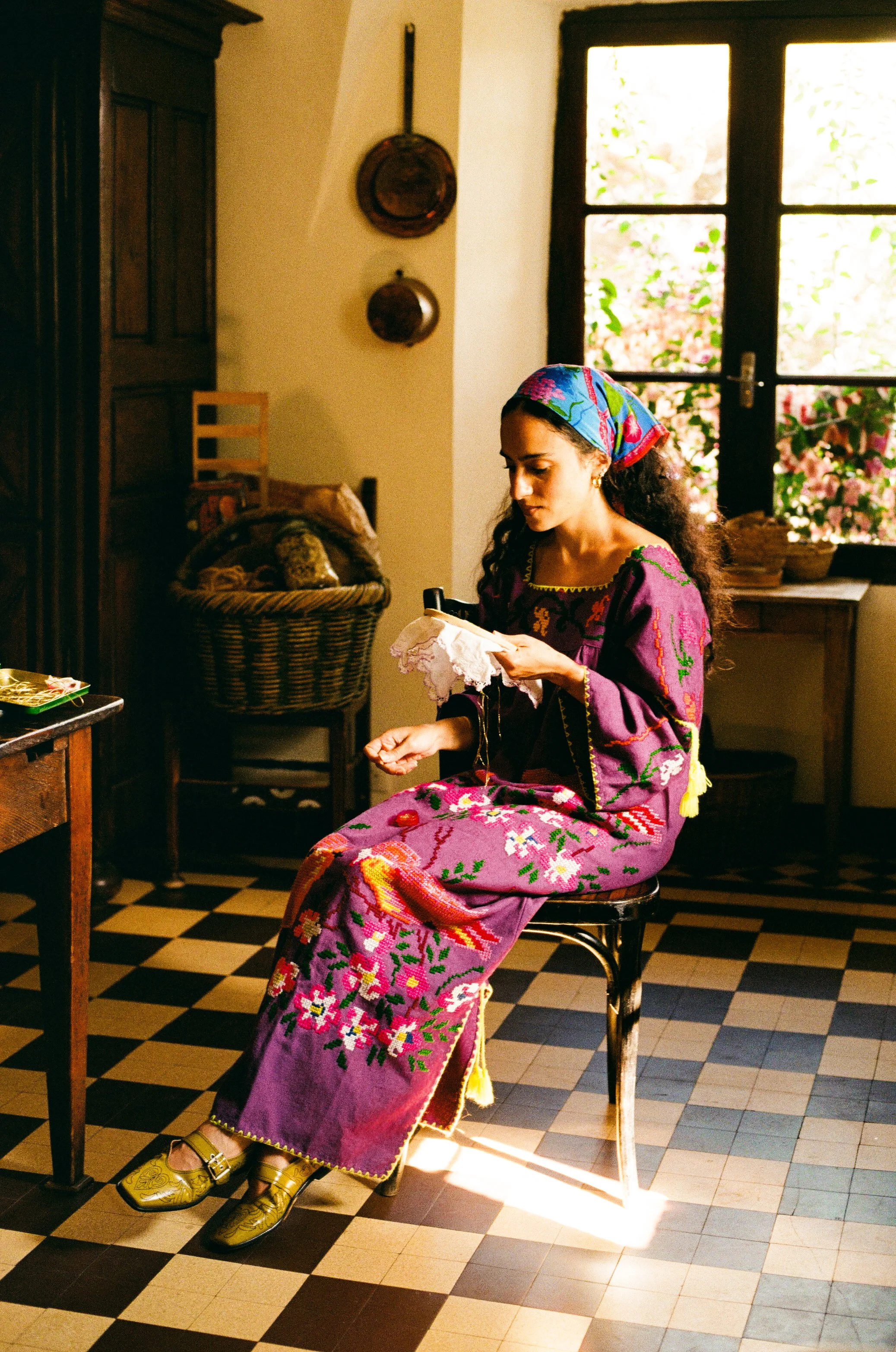 A young woman with curly hair, wearing a colorful headscarf, long embroidered dress, and yellow shoes, sitting on a chair in a kitchen with checkered black and white floor, reading a book or magazine. Background includes a window with flowers outside