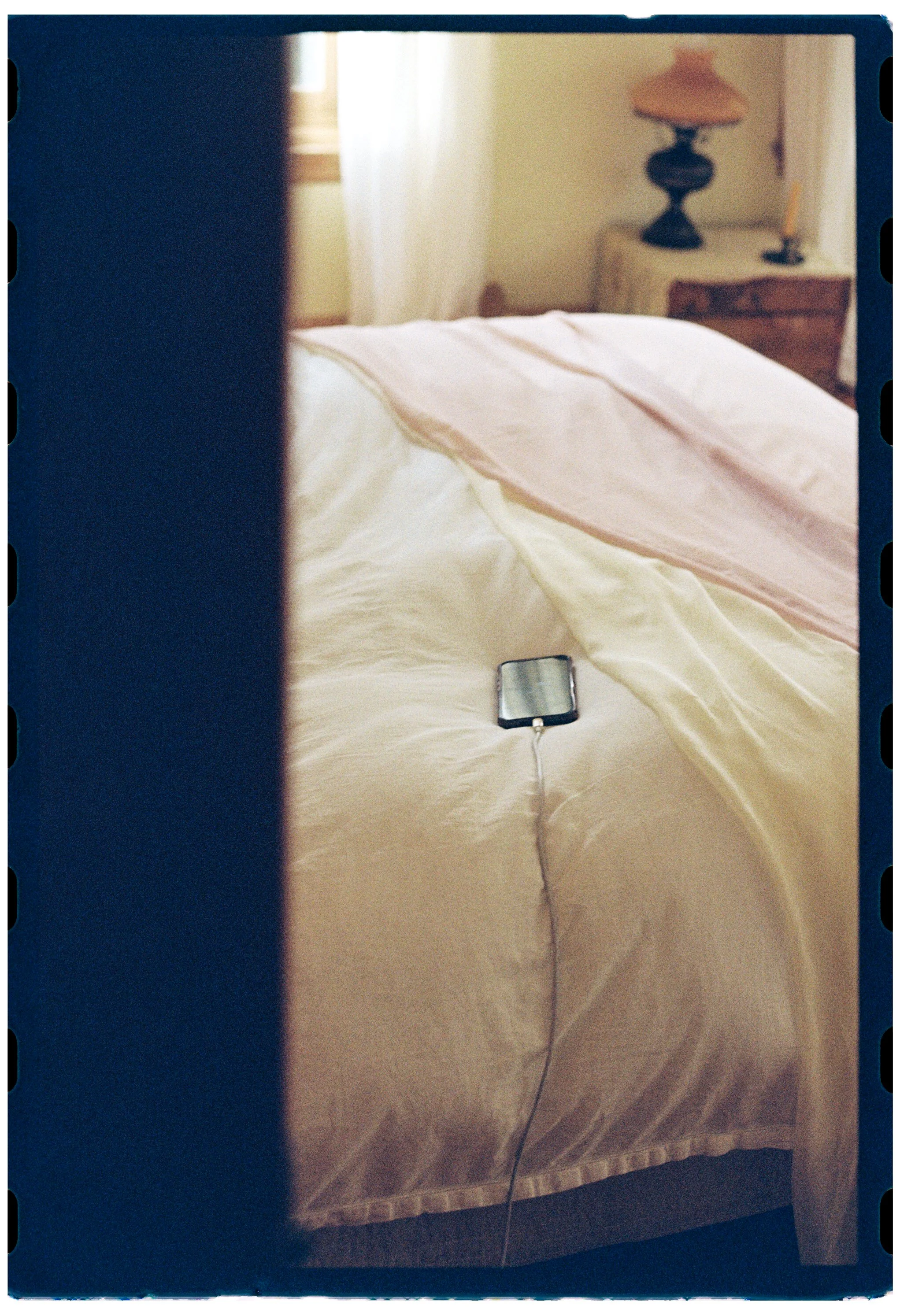 A bedroom with a bed covered in cream and pink sheets, a bedside table with a lamp, and a window with curtains, seen through a partially opened door.