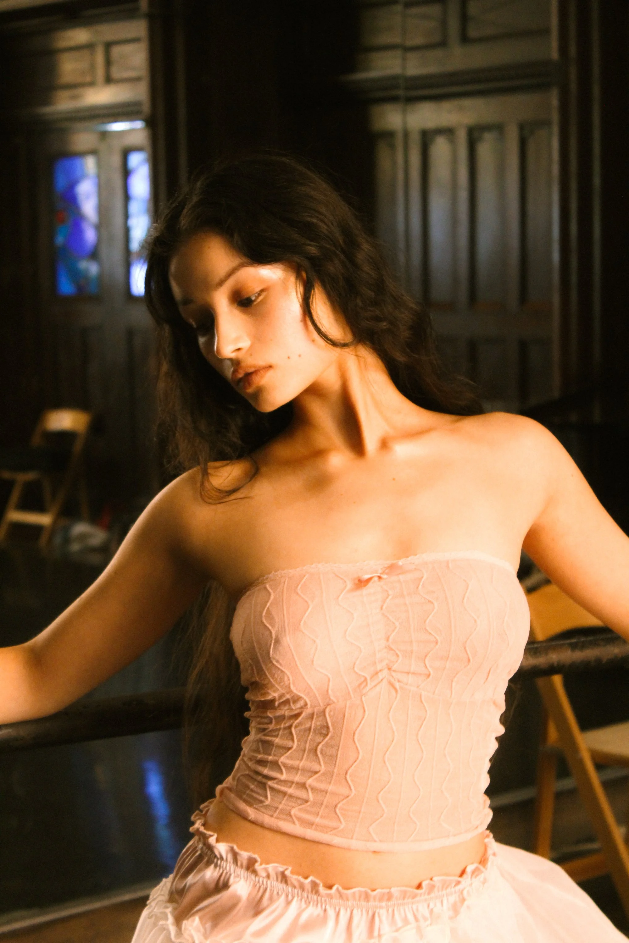 A woman with long dark hair looking down, wearing a strapless peach-colored dress with ruffles and a textured pattern, standing in a dimly lit room with dark wooden paneled walls and chairs.