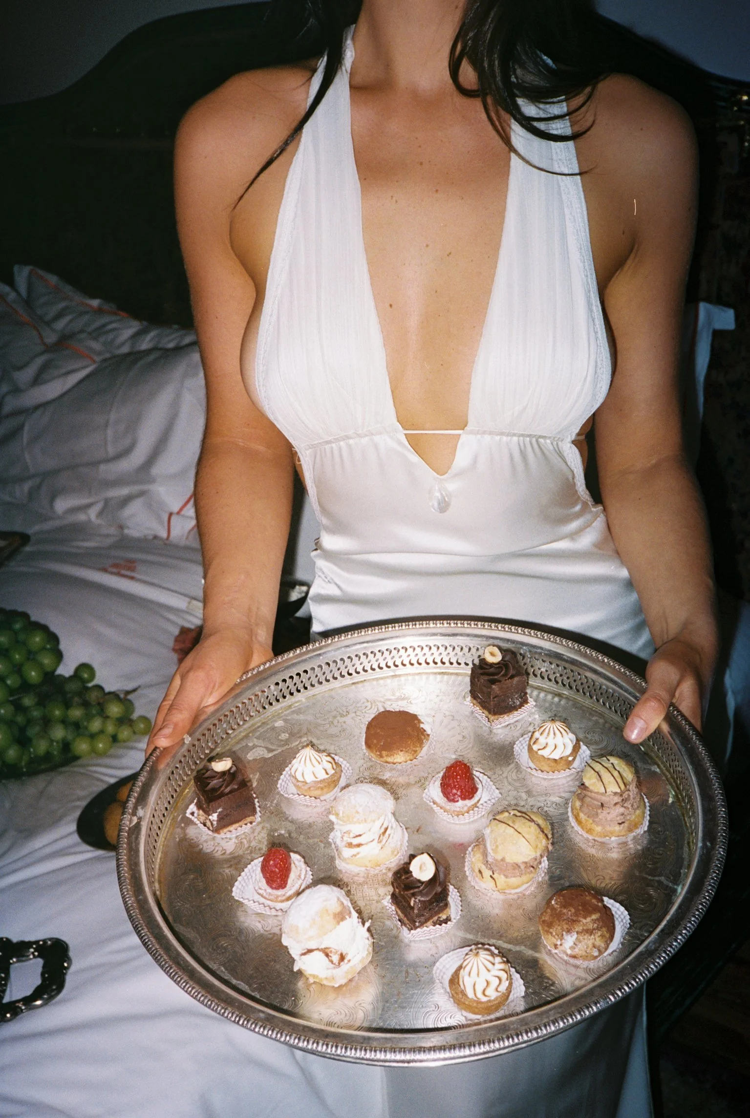 A woman in a white dress holding a silver tray with assorted small desserts, including cupcakes and pastries. There are green grapes visible on a bed nearby.