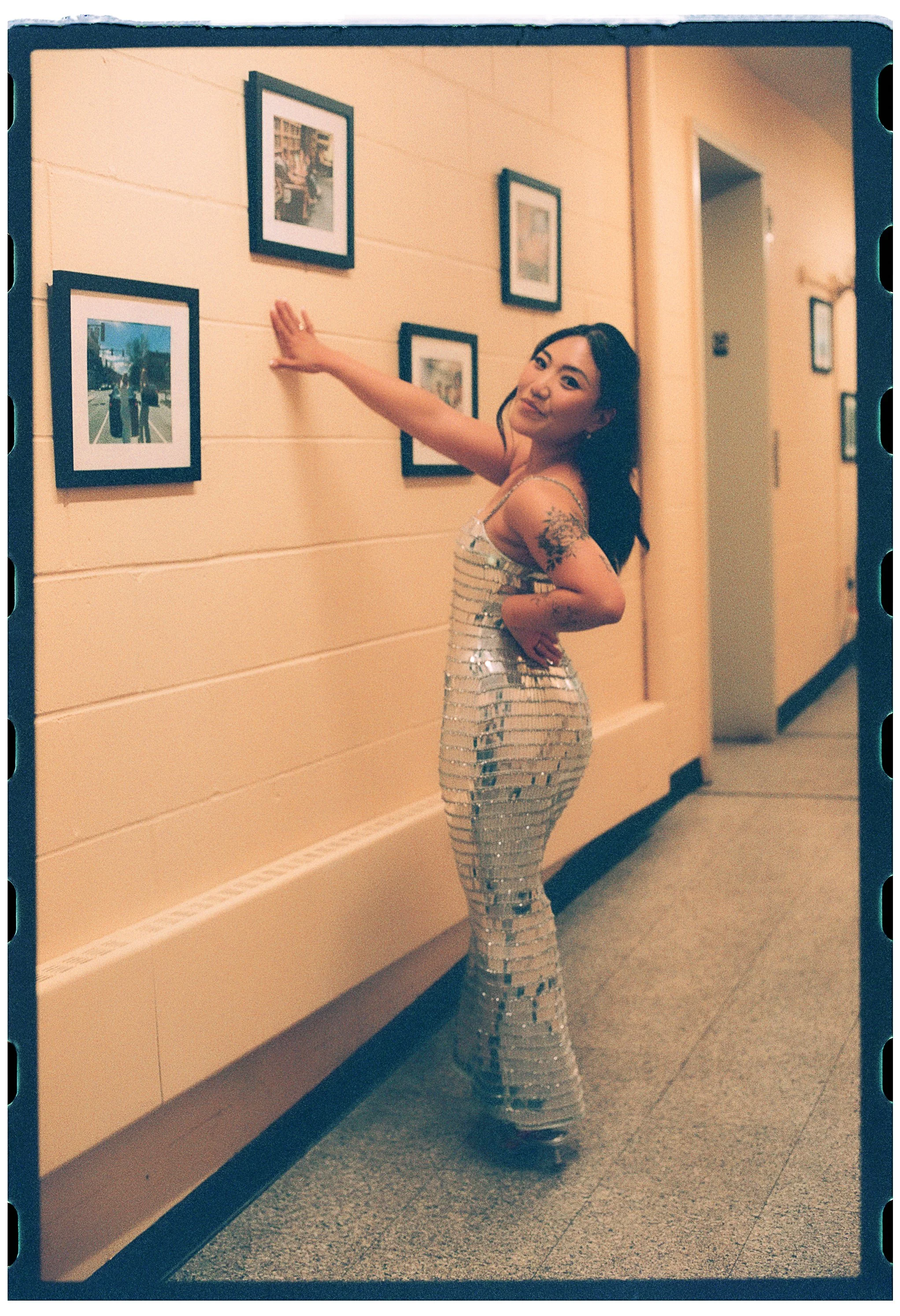 Woman in a shiny, sequined dress standing in an art gallery, looking at framed photographs on the wall.