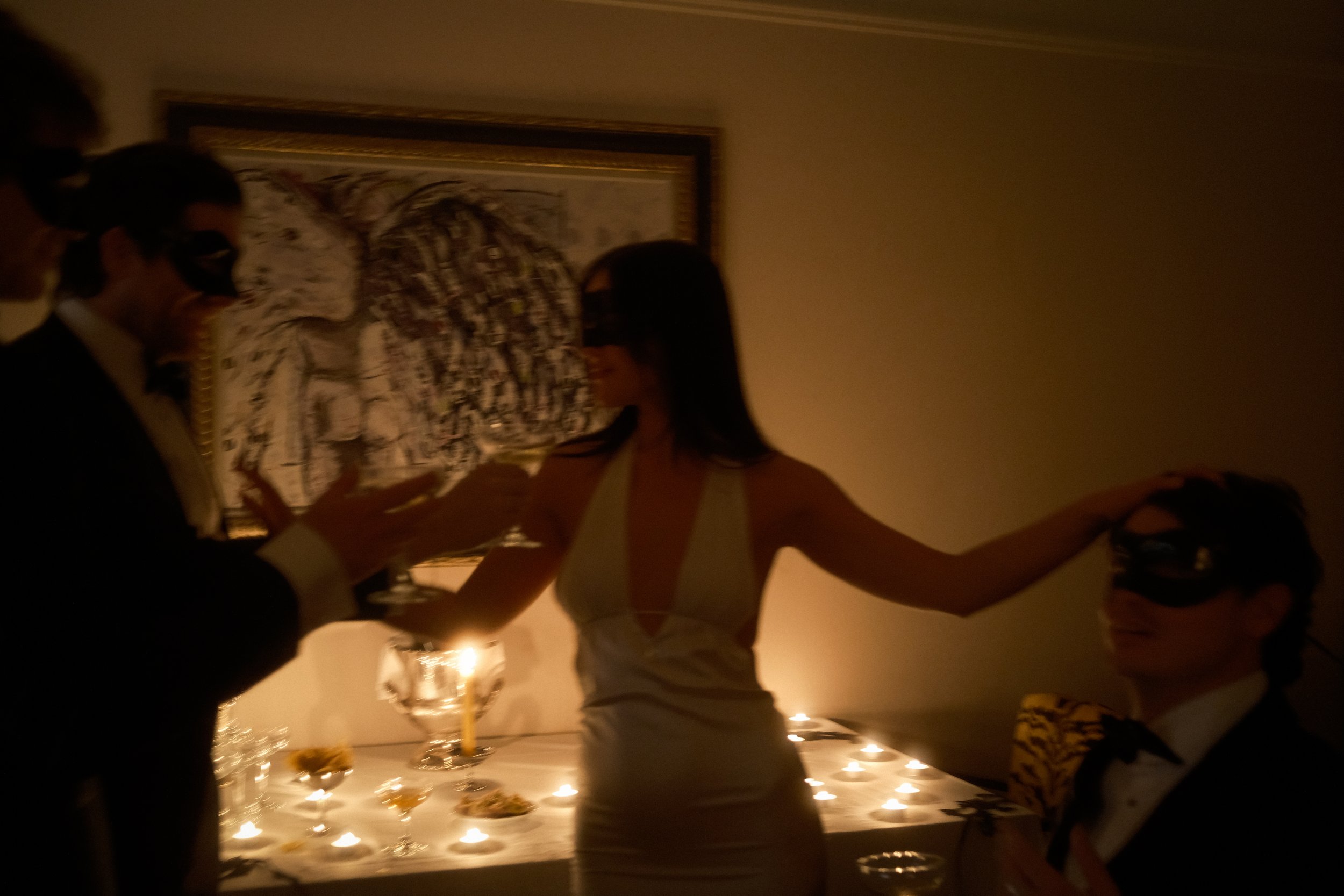 People holding hands in a circle at a party or gathering with dim lighting and candles, with a table of snacks or food in the background