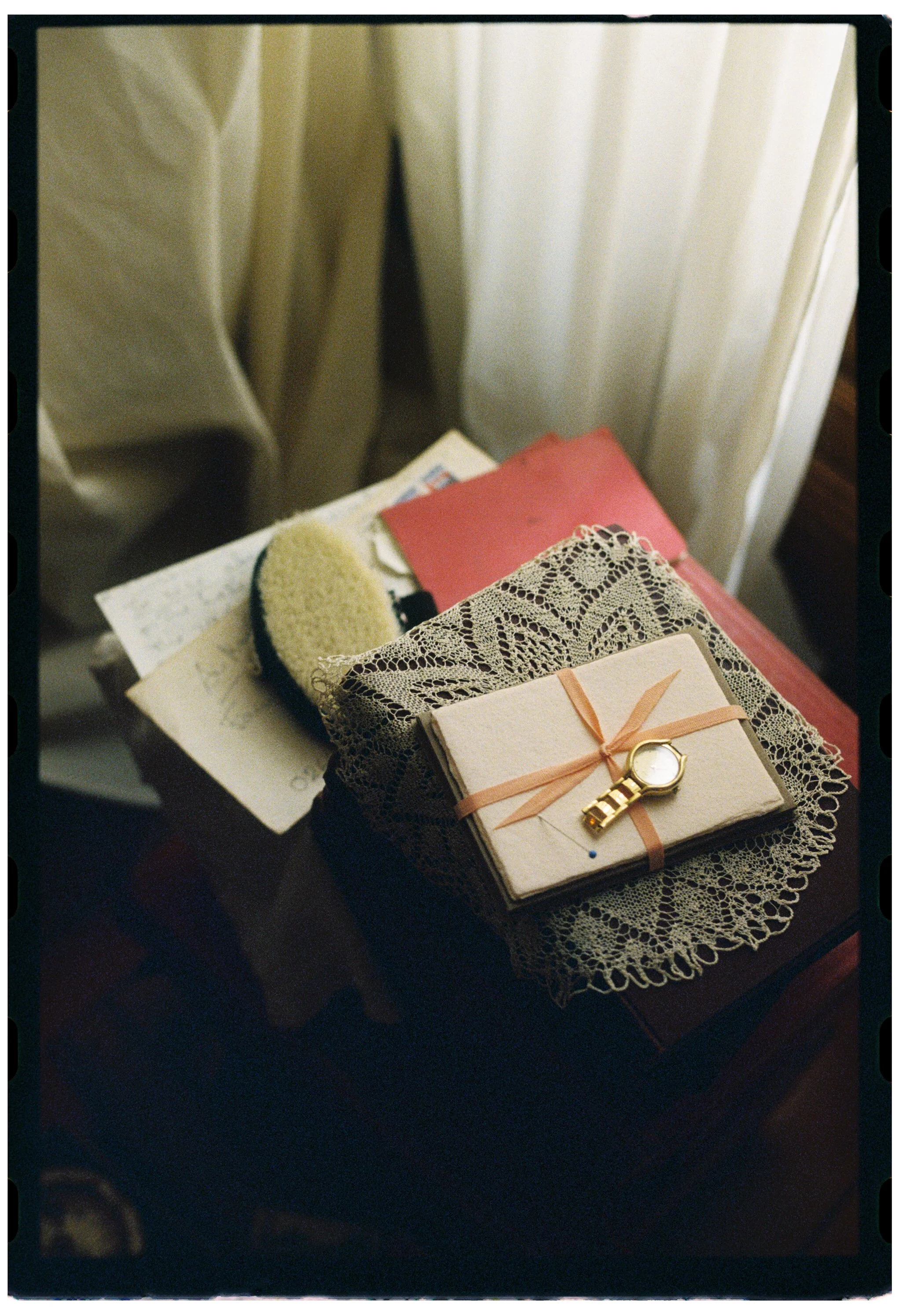 A table with a lace doily, a wrapped gift with a watch on top, a red notebook, and lace fabric. In the background, there are papers and a hairbrush on the table.