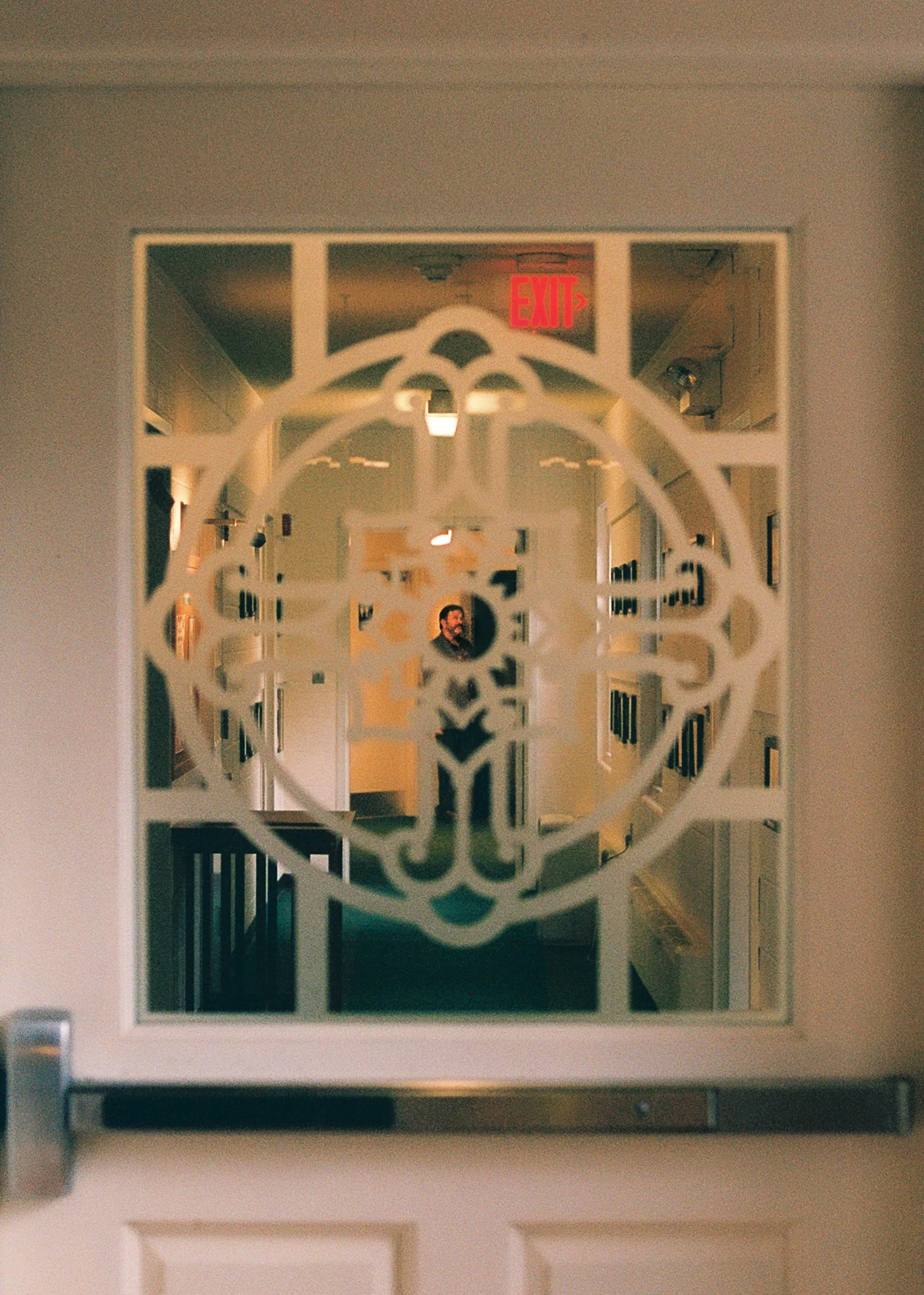 View through a decorative glass panel of a hallway with an illuminated red exit sign and a person with a beard walking in the distance.